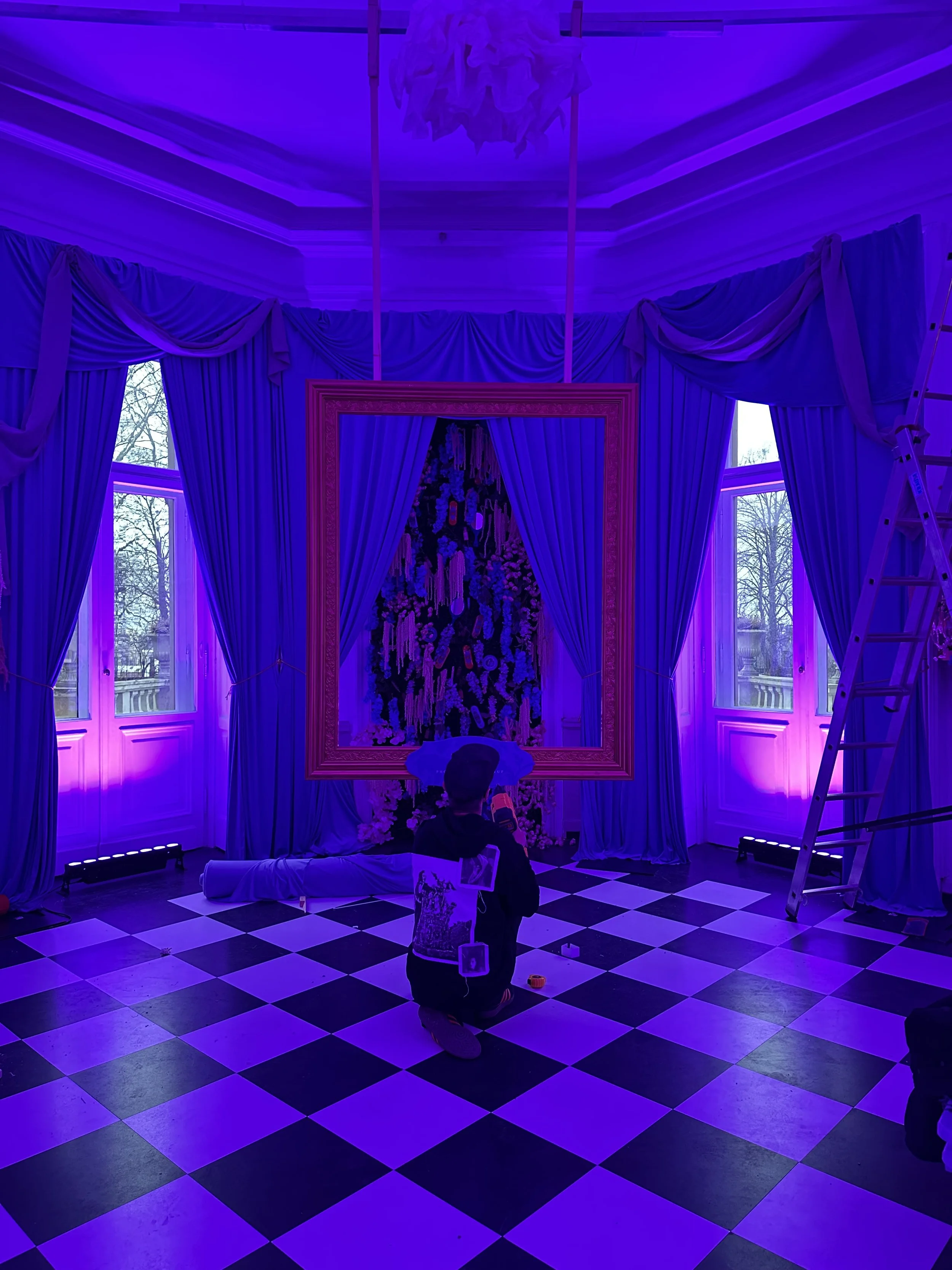 Room with checkered black and white floor, purple lighting, and large windows with curtains. A large ornate frame with floral decorations is centered, with a draped fabric at the base. A person is kneeling in front of the frame, holding a camera or p