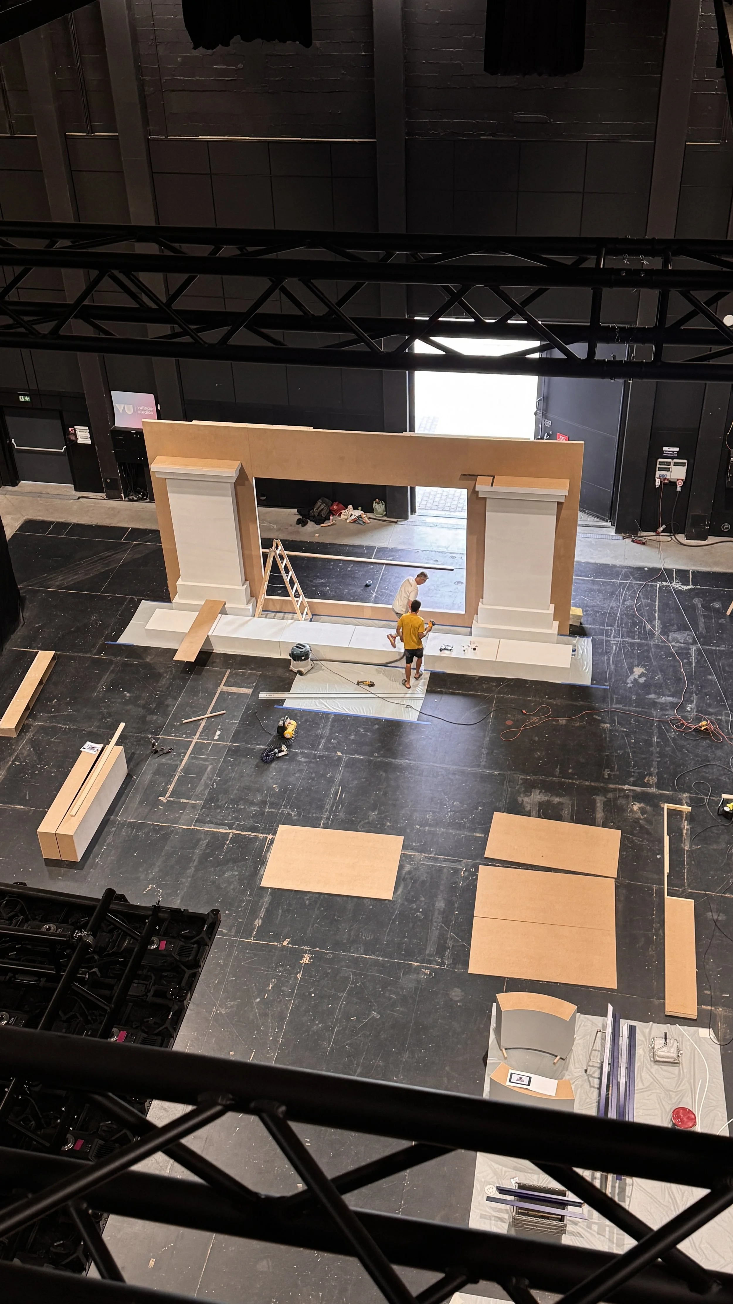 Two workers are constructing a large decorative frame or archway on a black floor with various tools and materials around them.