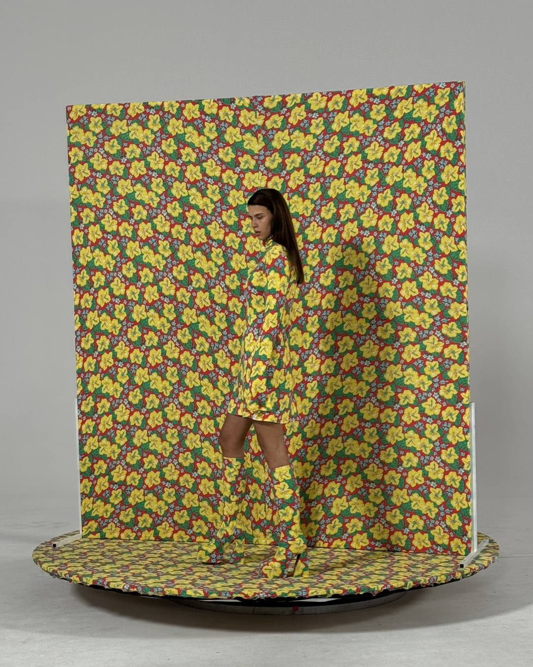 Woman in a floral yellow and red dress and matching boots standing on a circular platform with a floral backdrop.