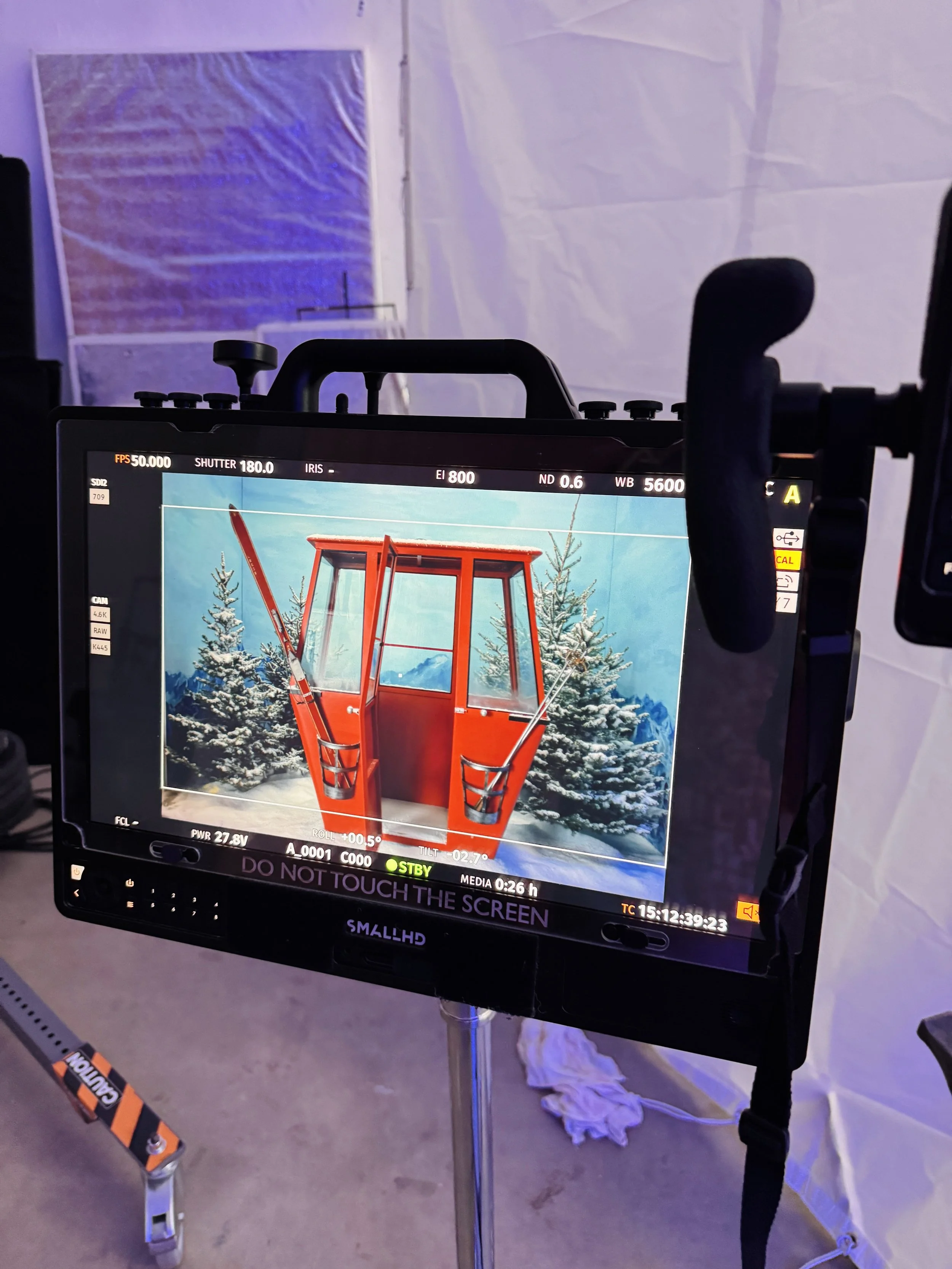 Close-up of a camera monitor showing a digital image of a red snowplow blade on a snowy landscape with pine trees in the background.