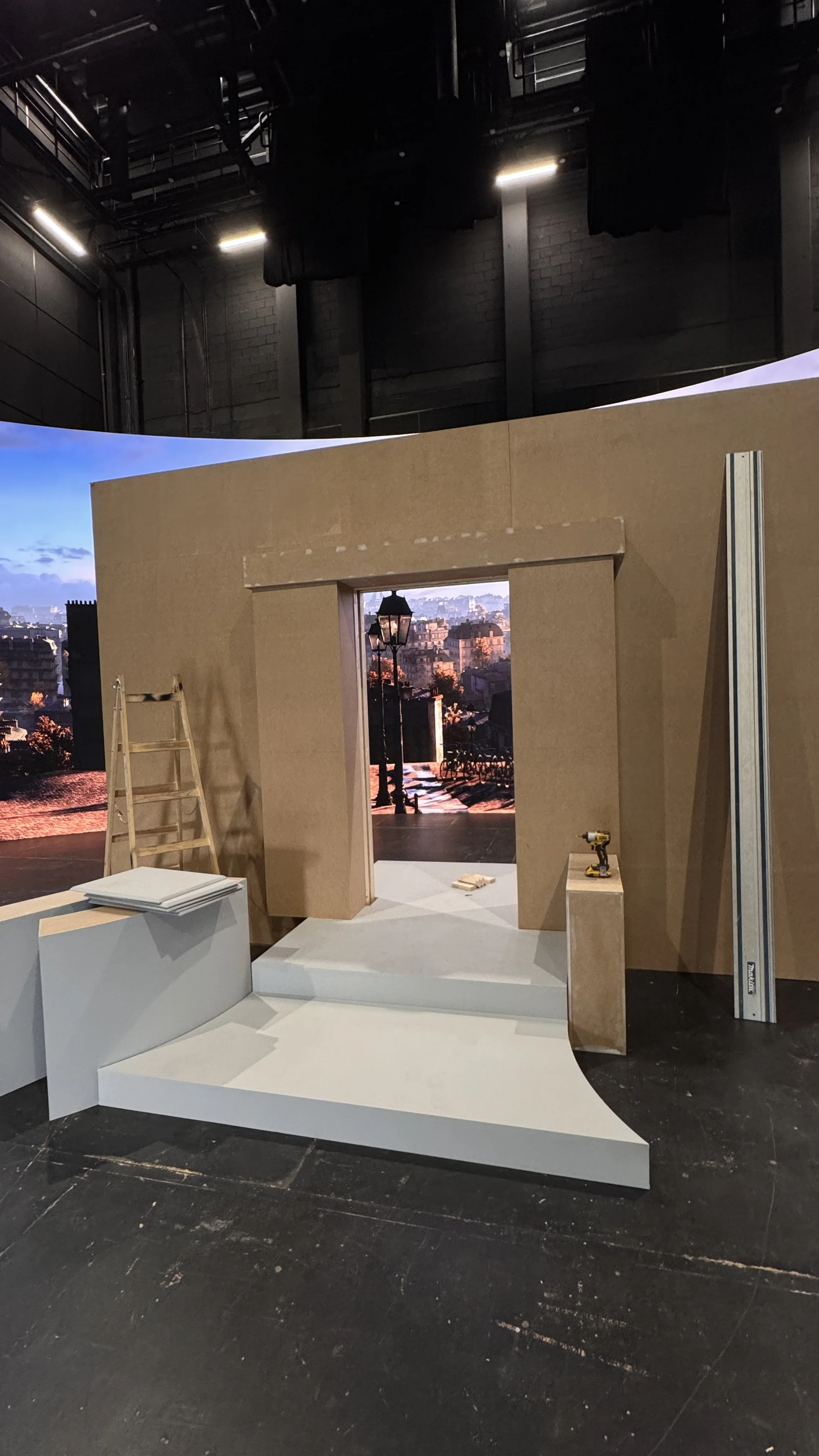 Stage setup in progress with a large, curved backdrop showing a cityscape at sunset, a wooden wall with a doorway, a ladder, a small white platform, and tools on the floor.