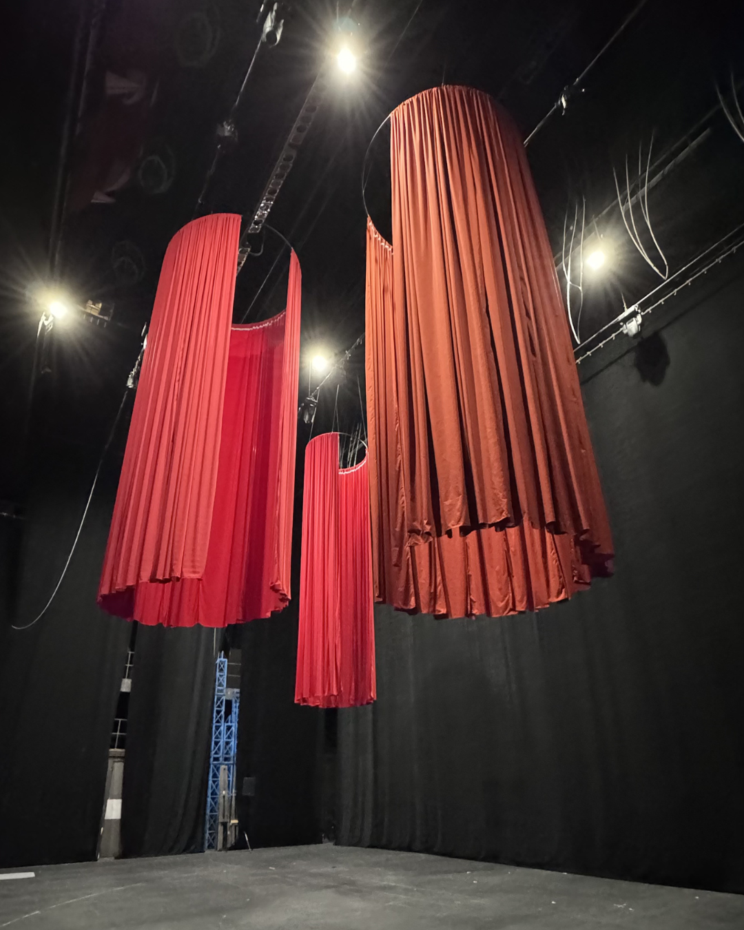 Three large red theater curtains hanging from the ceiling in a dark stage, illuminated by spotlights.