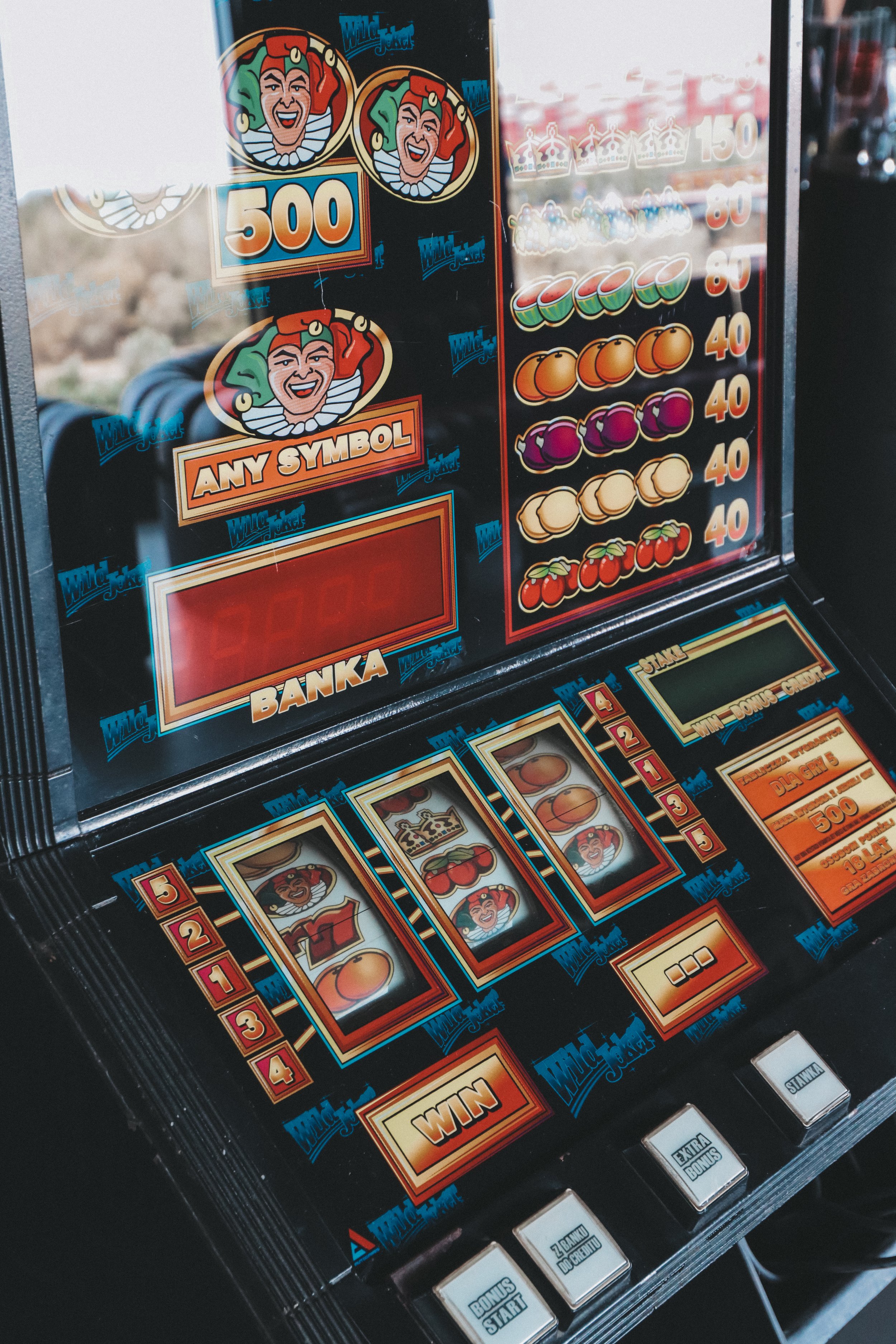 Close-up of a vintage pinball machine featuring a jester mascot with a red and green hat and a wide smile. The machine displays various colorful symbols and numerical scores, including a large "500" point marker. The lower section shows buttons label