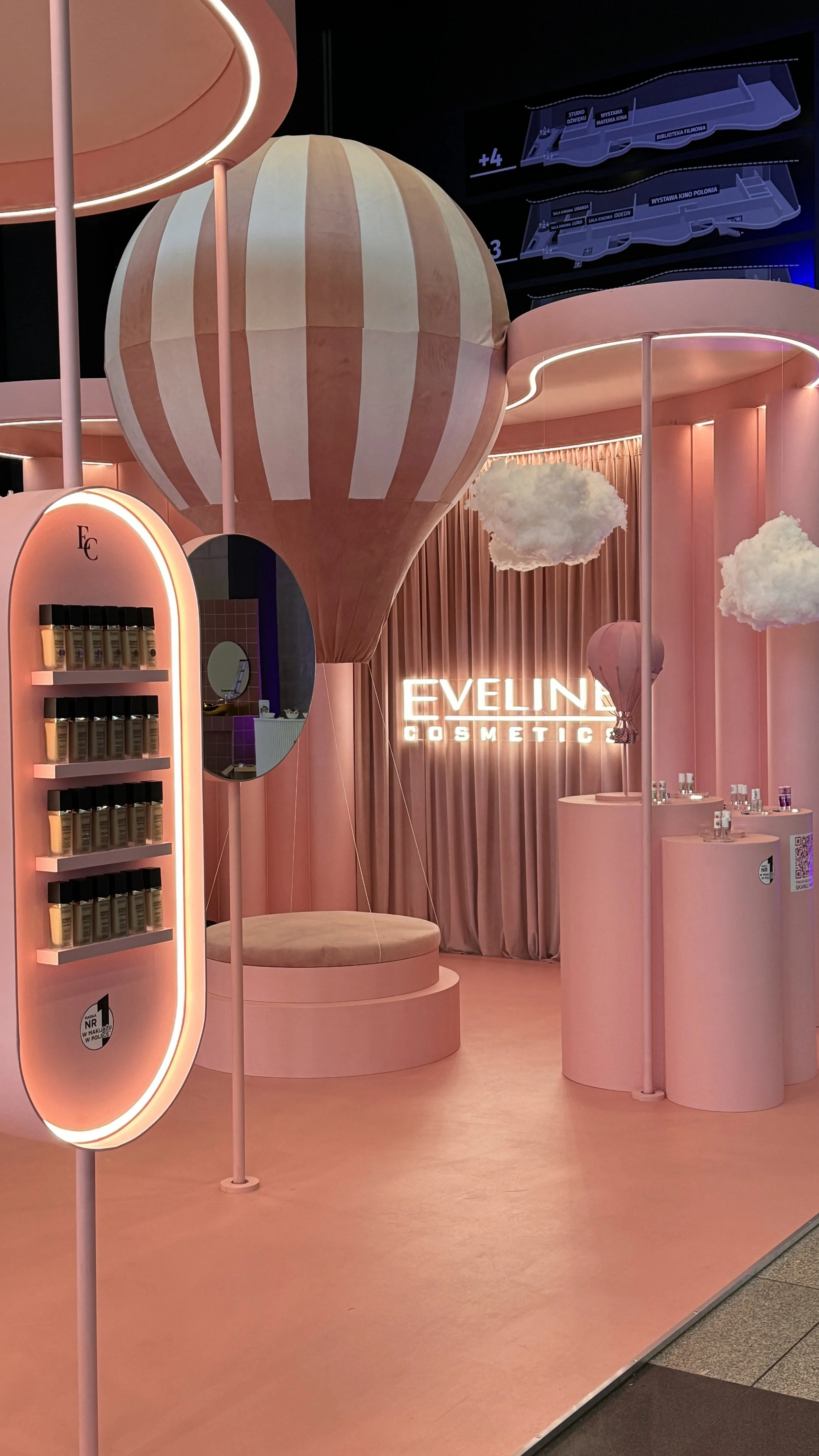 The image shows a colorful display booth with a hot air balloon theme for Eveline Cosmetics. The booth has pink and peach tones, decorative clouds, and the Eveline Cosmetics sign illuminated in pink. There are shelves with products, a mirror, and sea