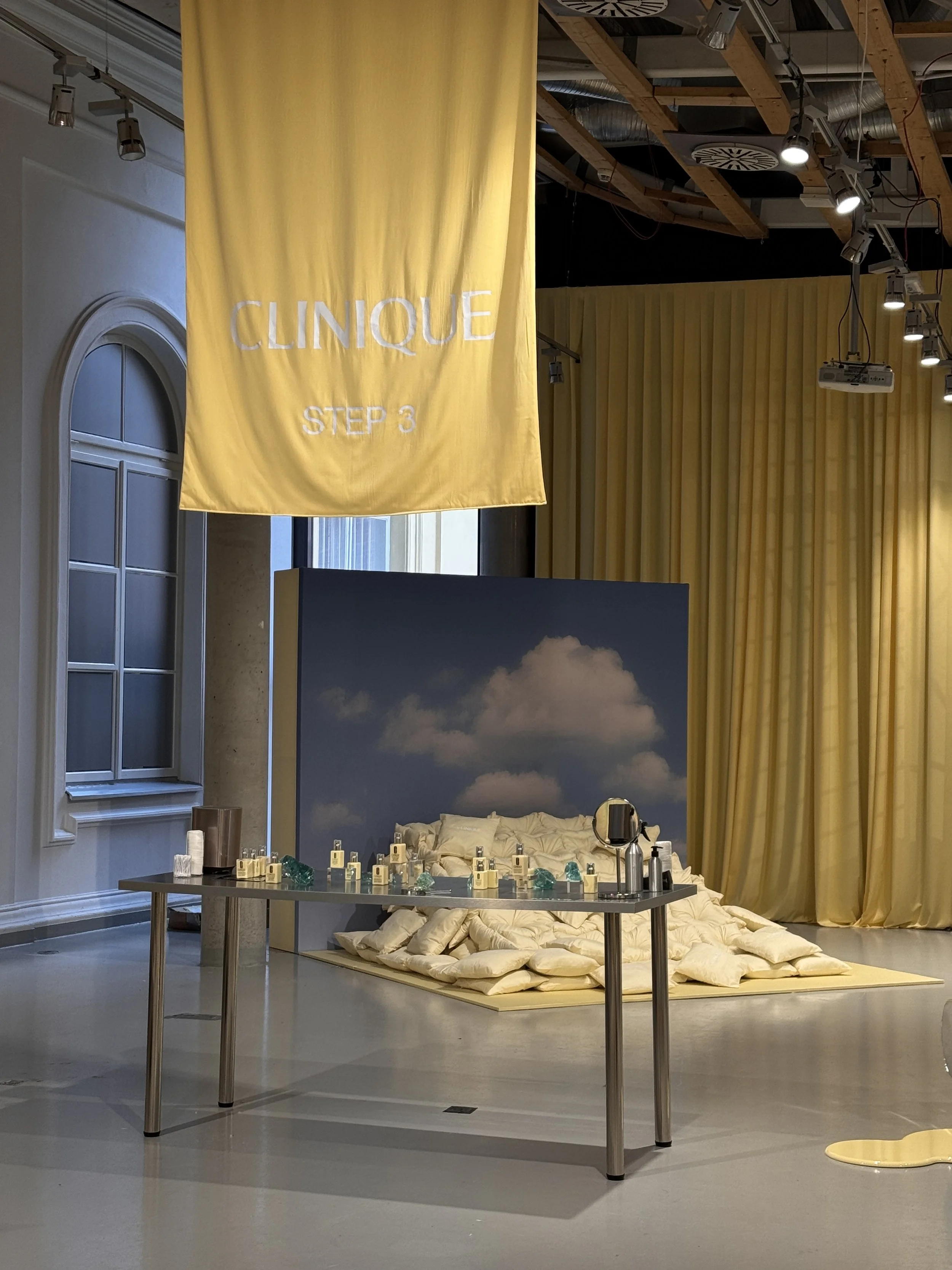 Interior of a room with a yellow banner reading 'CLINIQUE STEP 3'. There is a table with skincare products and green glass decorations, a bed with cushions and pillows, yellow curtains, and a high ceiling with exposed wooden beams and hanging lights.