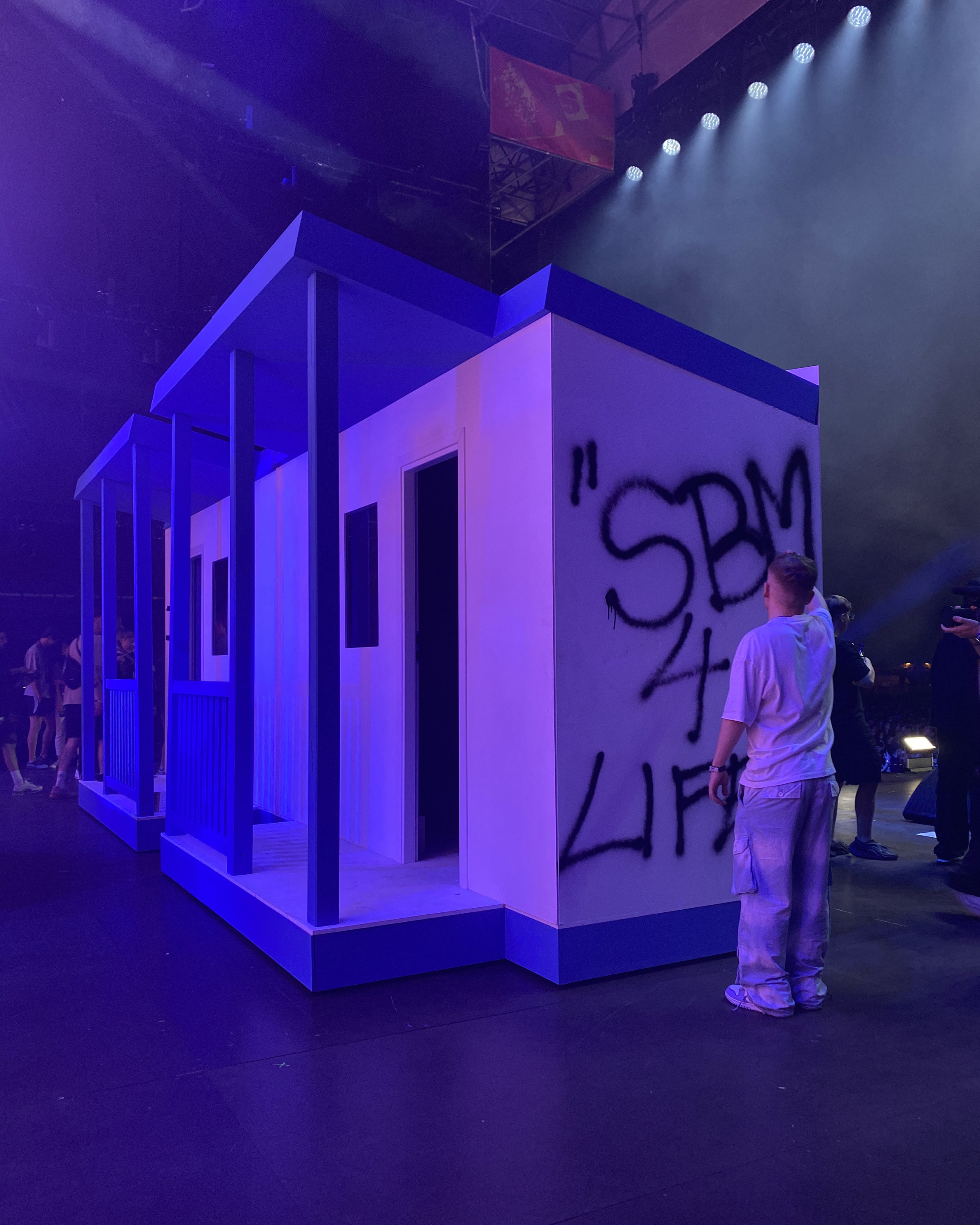 Indoor event with a modern, minimalist white structure booth illuminated by purple and blue lighting, with attendees standing and walking around, some of whom are wearing white clothing, and graffiti on the kiosk side with the words 'SPRE...'