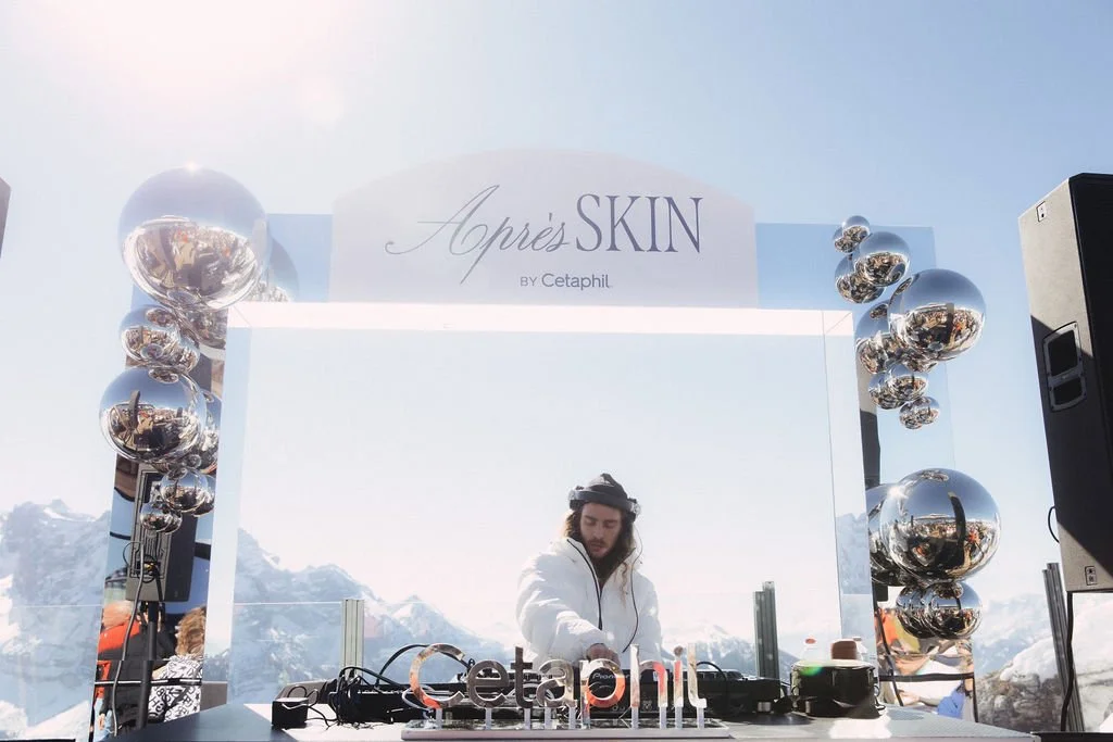 A male DJ in a white jacket and headphones performing at an outdoor event set against snowy mountains, with a backdrop featuring a sign for Après SKIN by Cetaphil and reflective chrome balloons.