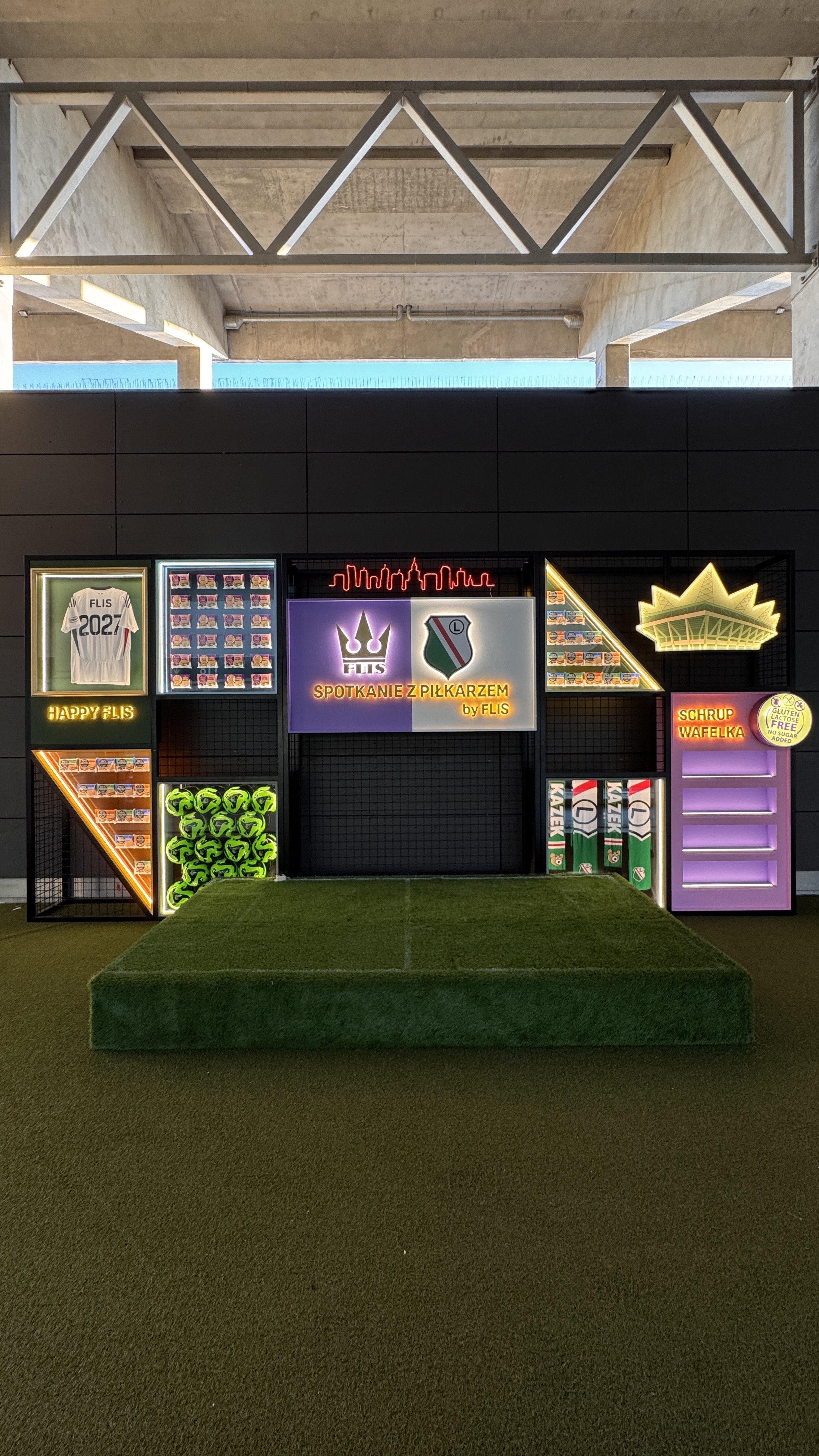Neon sign with a crown, a football helmet, and a badge with the text "SPOTKANIE Z PIŁKARZEM by FLIS". Additional signs include a jersey with the number 202 and the name FLIS, a crown, and a purple shelf with empty space. Green soccer balls are displa