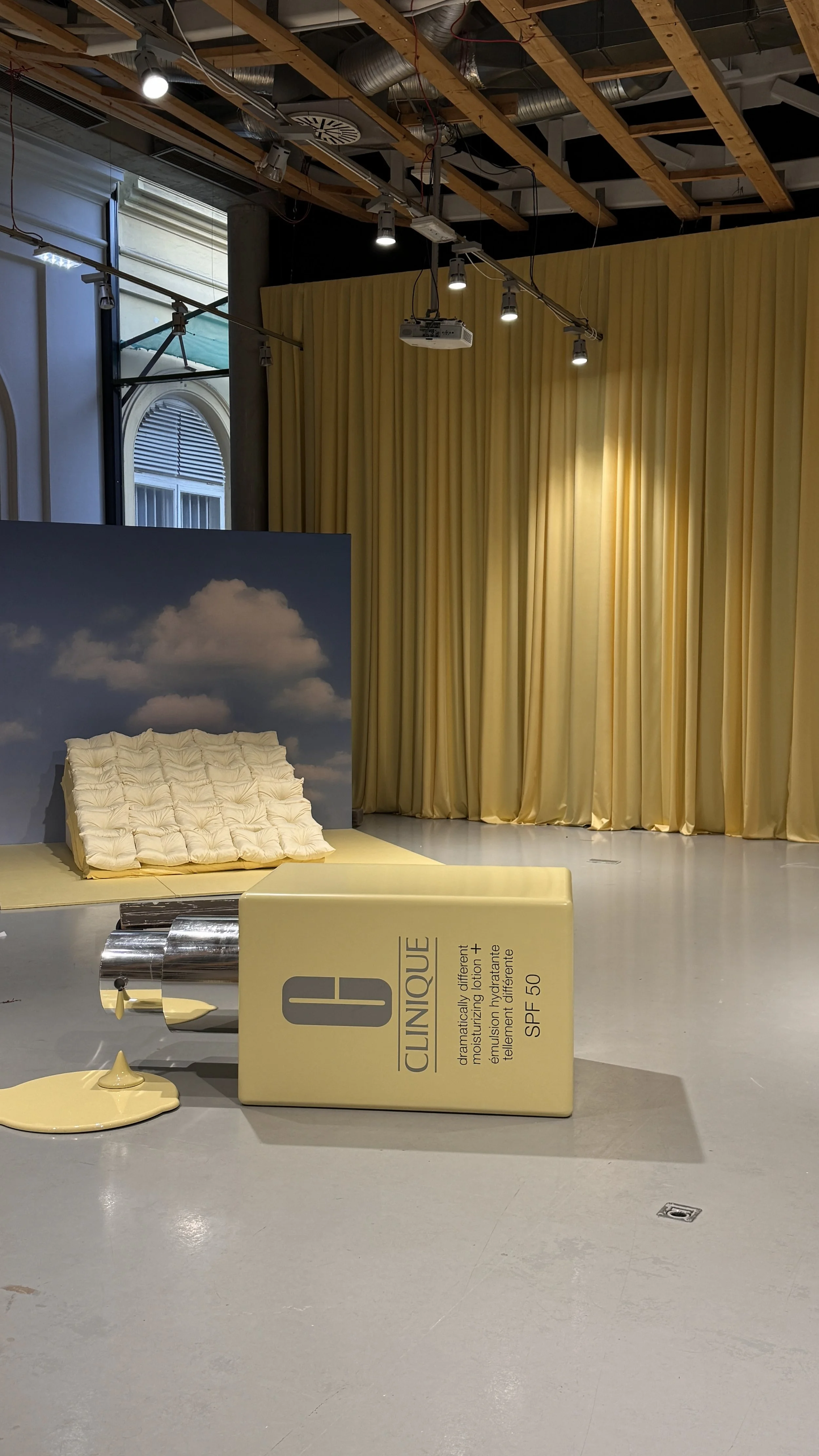 A room with a yellow curtain, a large cushion on the floor, a backdrop of a cloudy sky, and a large Clinique skincare product on display.
