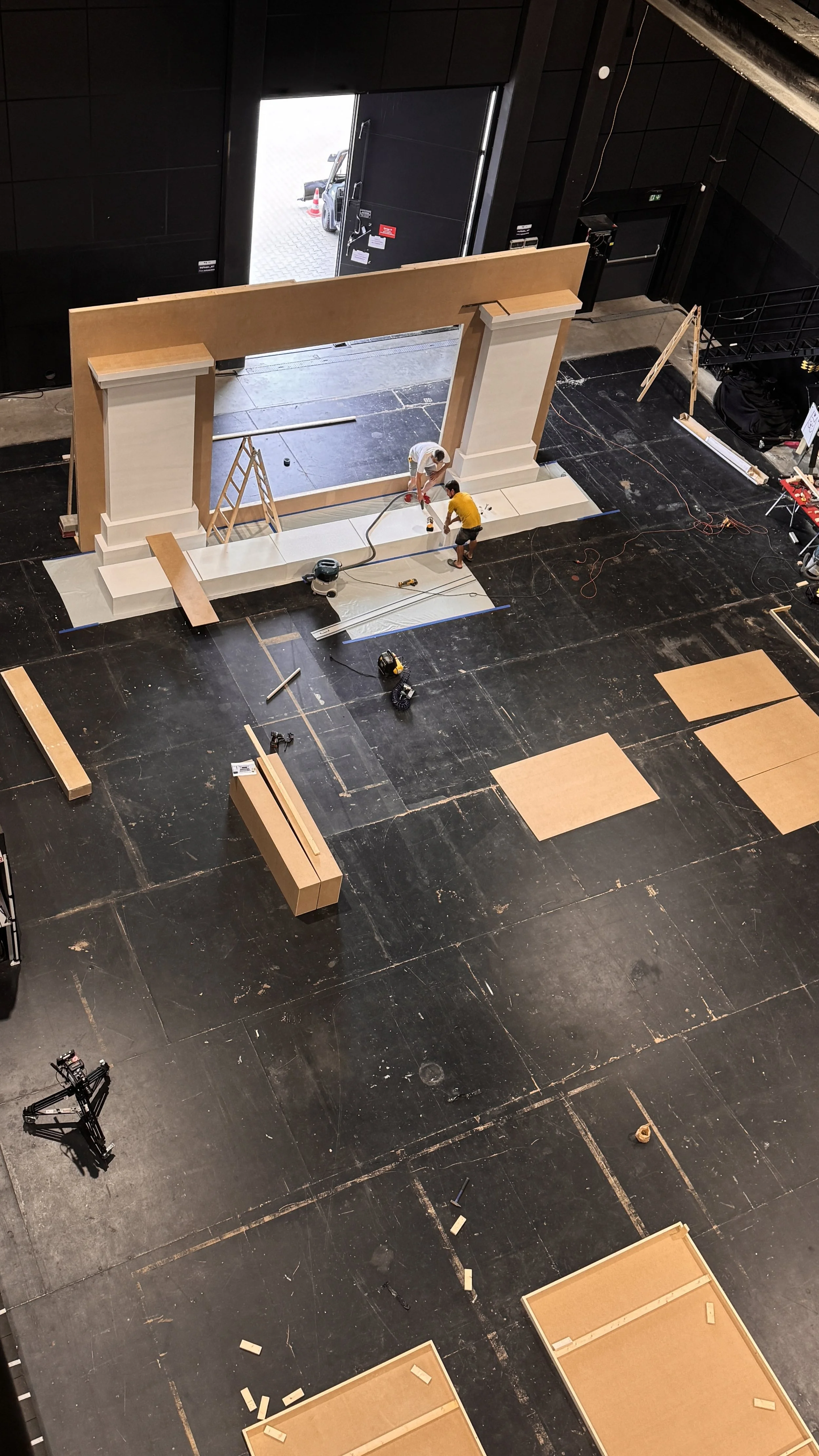 Two people working on a construction project indoors, assembling large white panels in front of a black wall and large open door with an outside view of a vehicle and traffic cones.