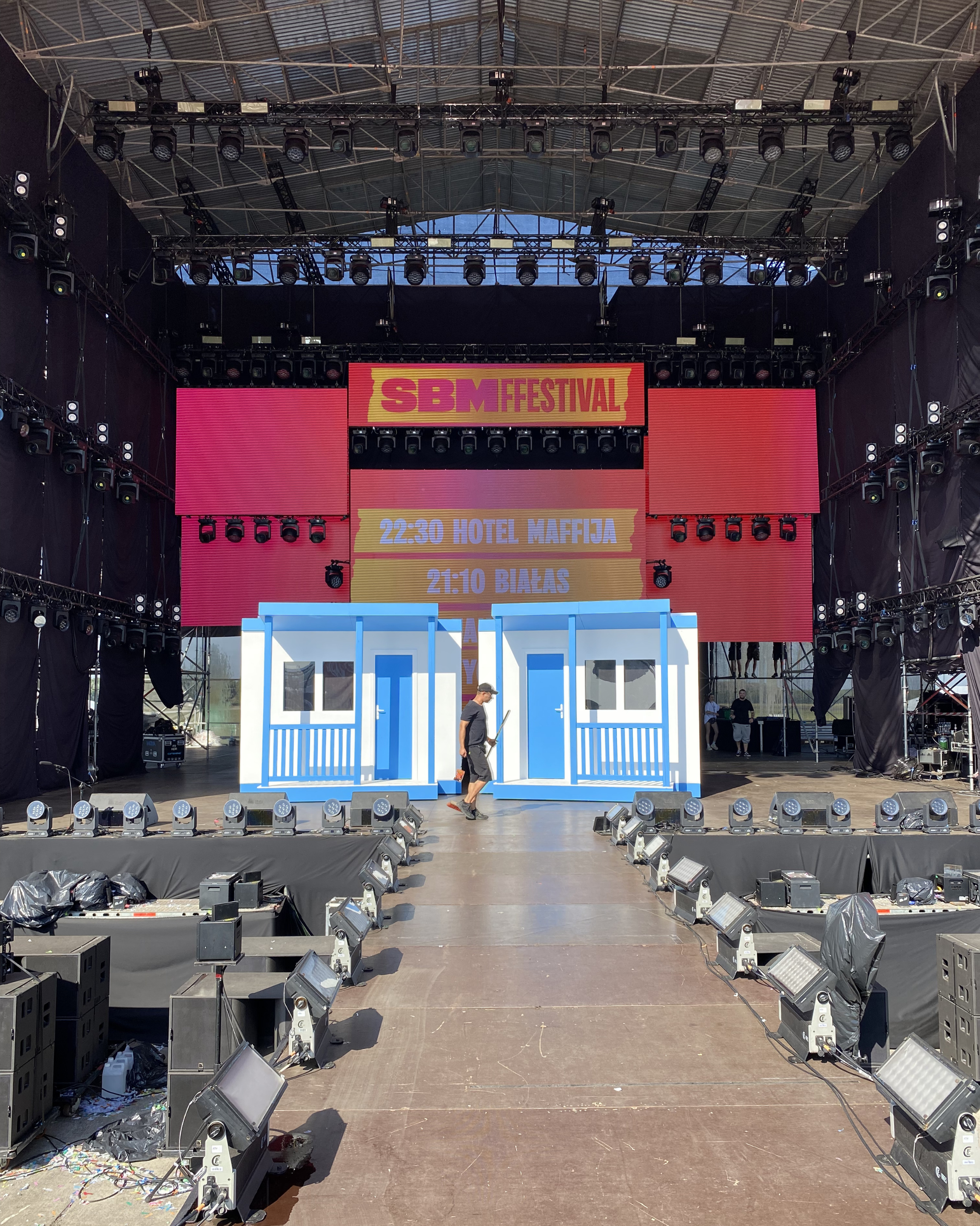Large outdoor stage setup with lighting and sound equipment, including a central screen displaying an event schedule for SBM Festival. There are no performers or audience visible in the image.