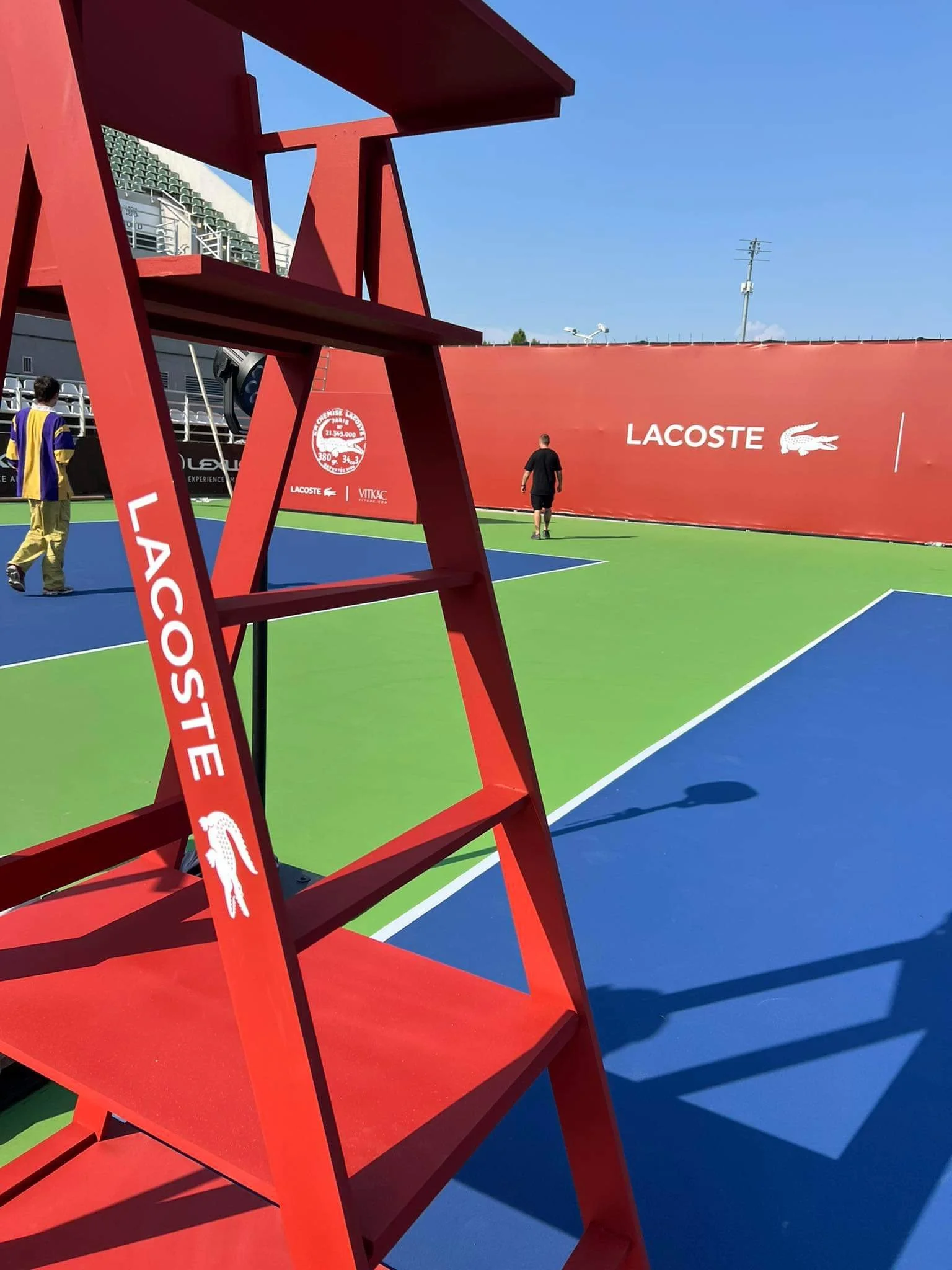 Red Lacoste branded umpire chair on a blue tennis court with a bright orange wall displaying Lacoste and KIAK logos, a small patch of green, and a clear blue sky.