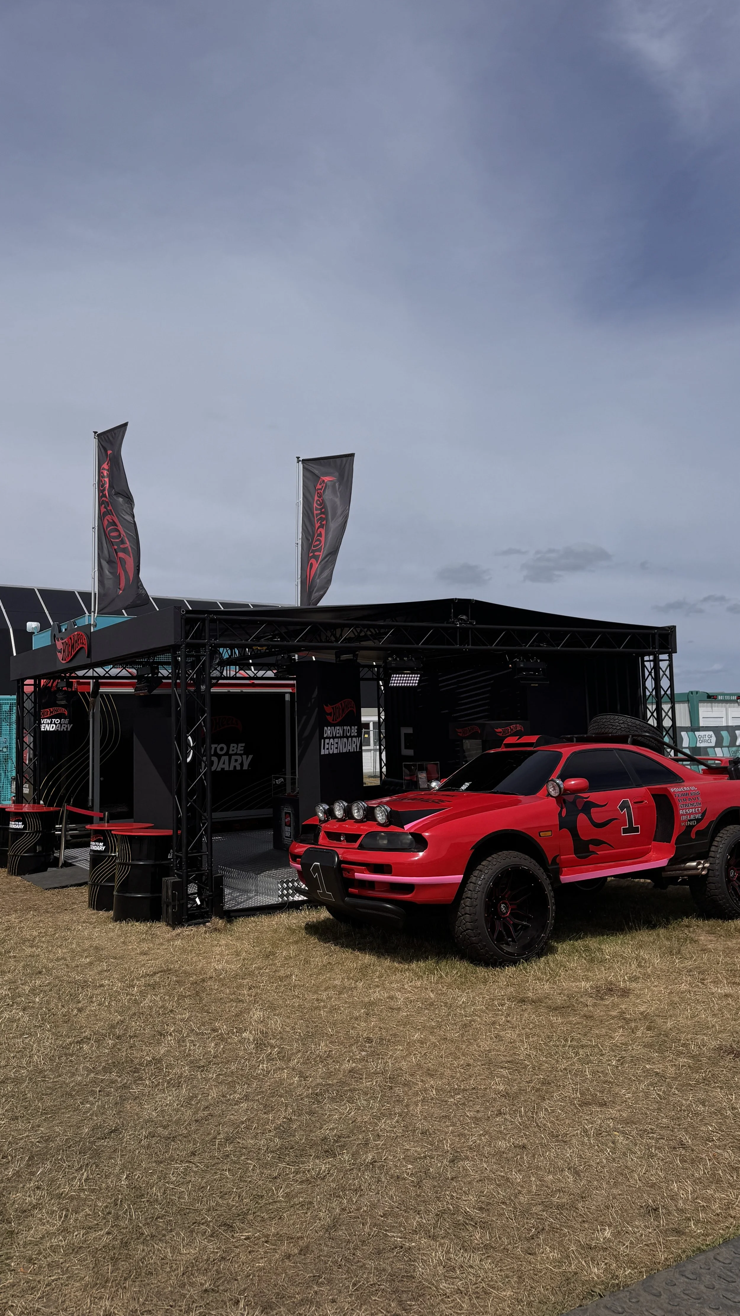 Red vintage race car parked in front of a black racing display tent with flags, under a blue sky with some clouds.