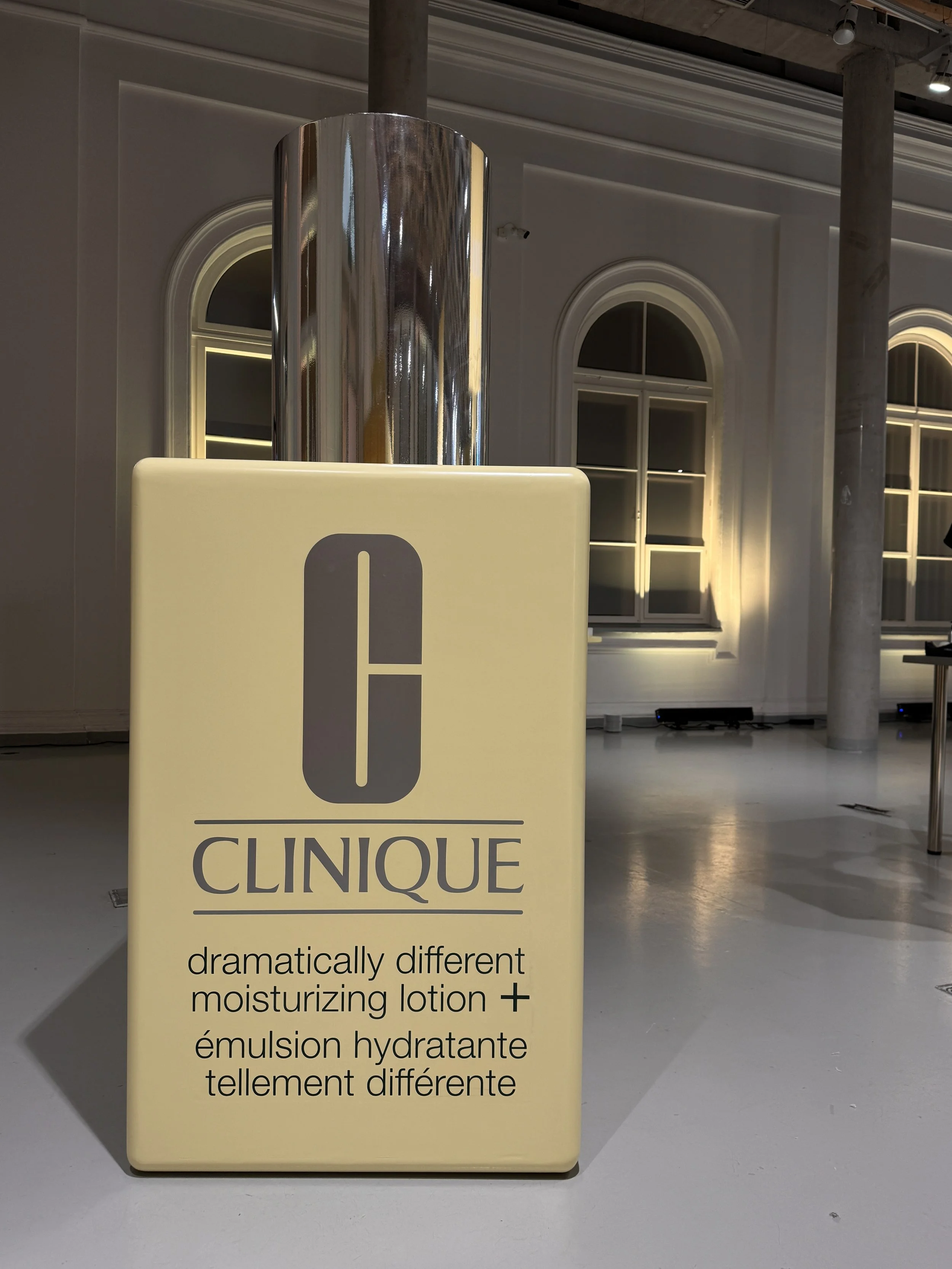 A beige sign with the word "CLINIQUE" and description of a moisturizing lotion in English and French, displayed in a well-lit modern interior with windows and columns.