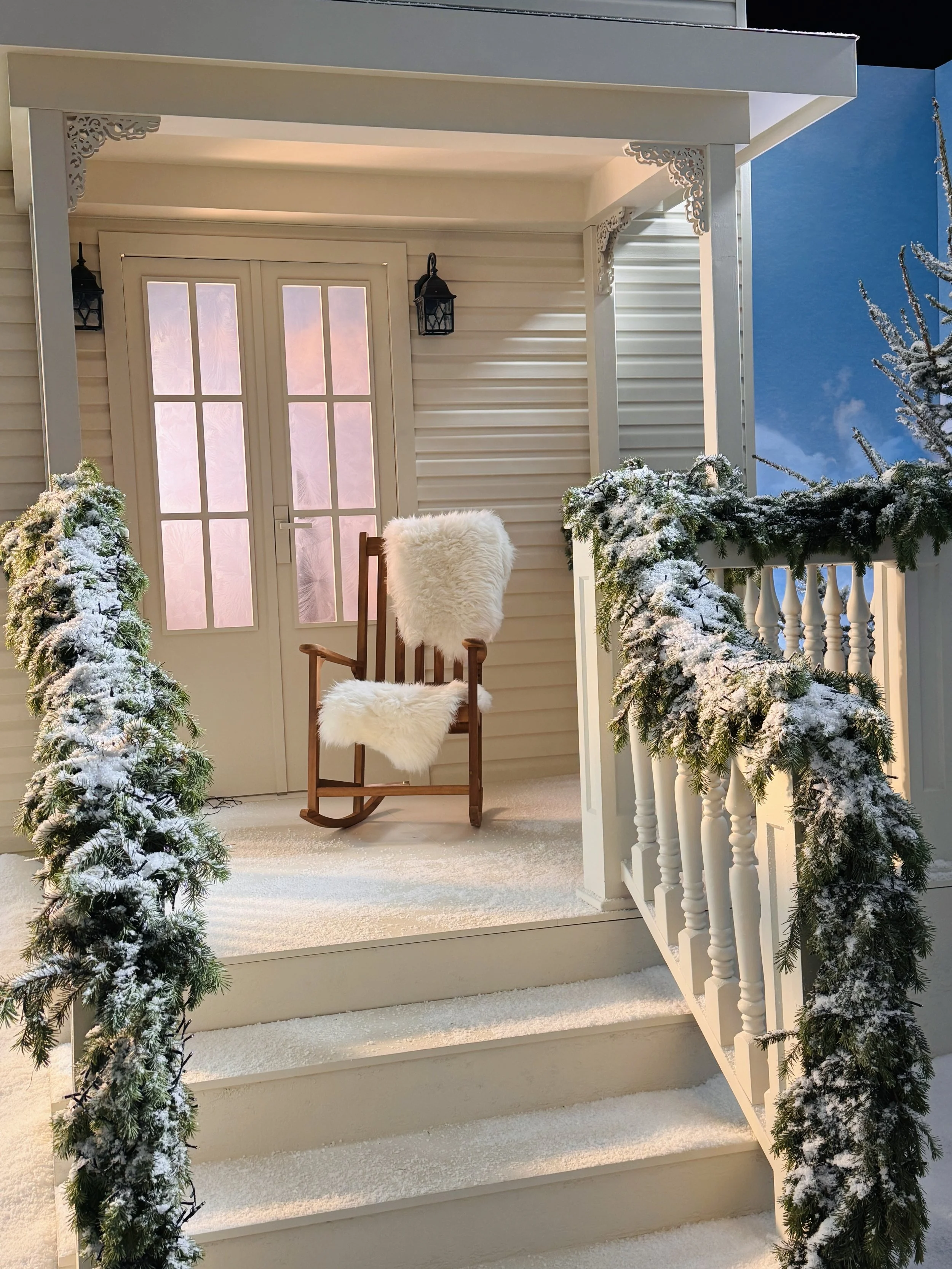 A snowy porch with a wooden rocking chair covered with a white fur throw, surrounded by snow-dusted evergreen garlands, and decorated with two black lanterns on the wall.