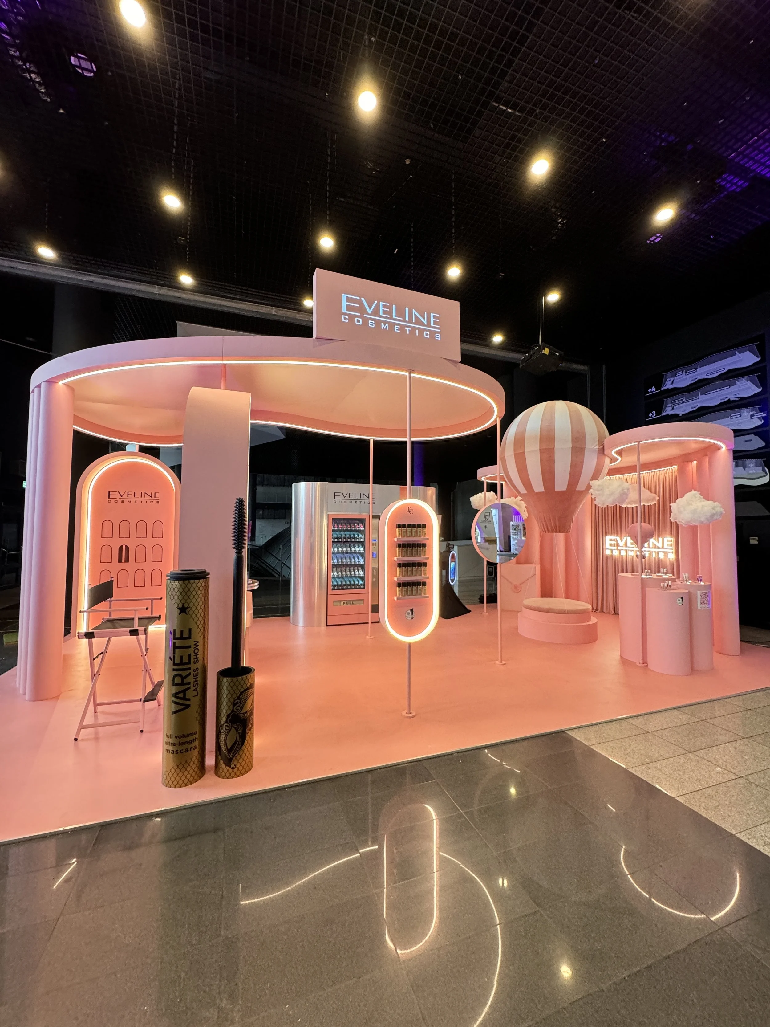 Pink-colored beauty booth with the sign 'Eveline Cosmetics' at an indoor event, featuring skincare and makeup products, illuminated with neon lights and decorative elements.