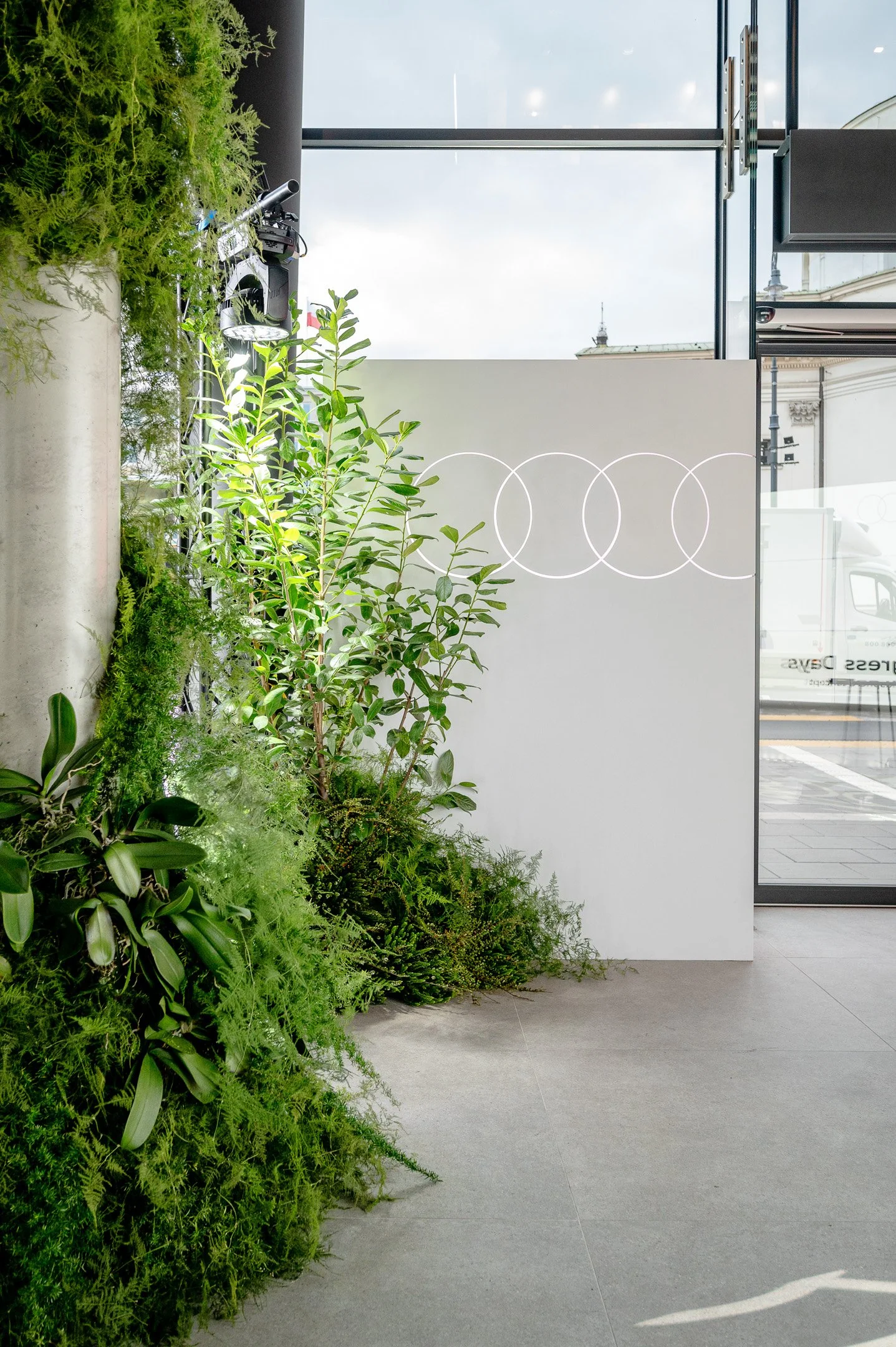 Green plants and foliage inside a modern building with large glass windows and a gray tiled floor.