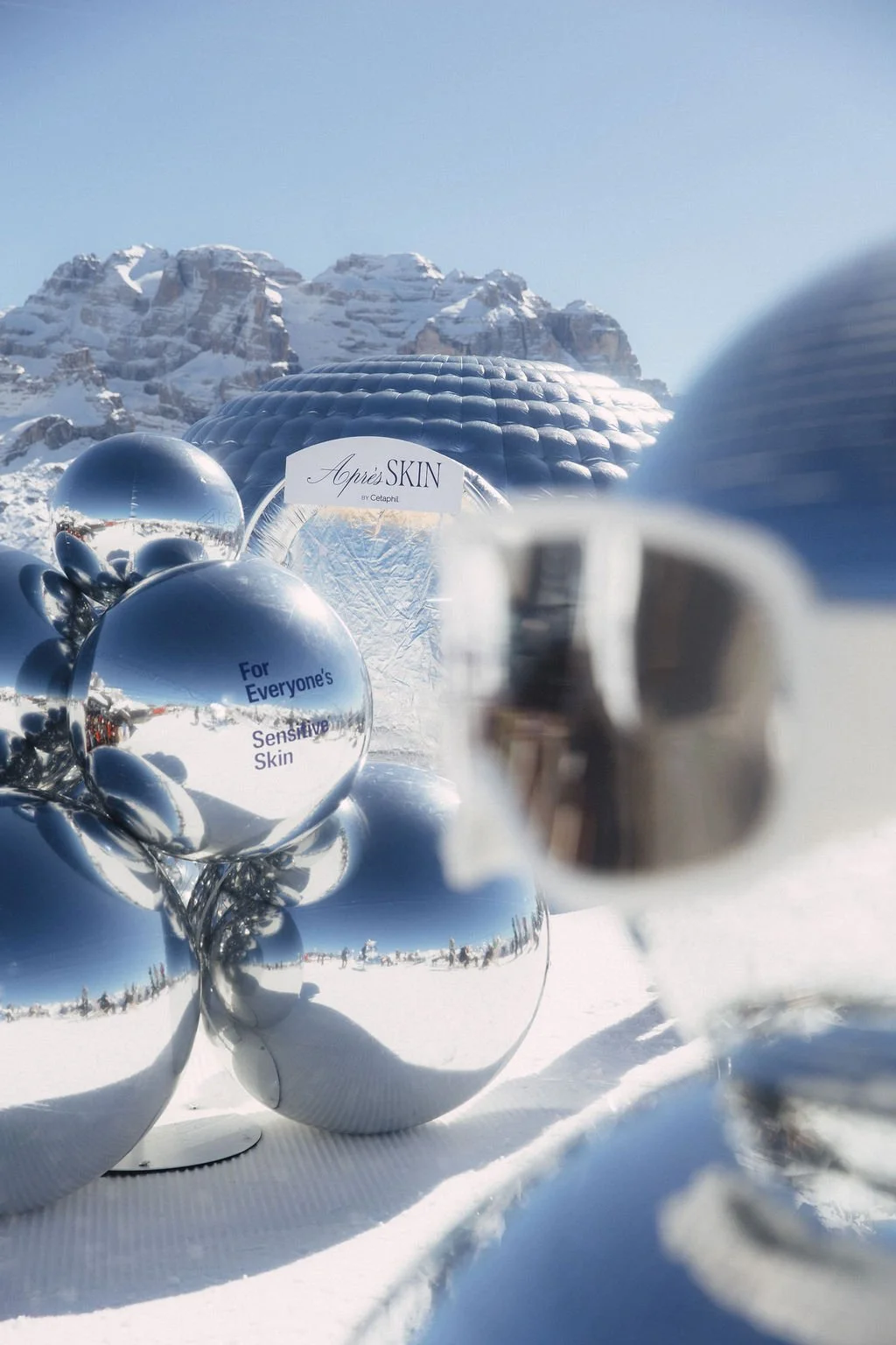 Reflection of a snowy mountain landscape in shiny silver spheres and a mirror-like dome, with a sign that reads, 'Après SKIN by Cetaphil' and a smaller sign that says, 'For Everyone's Sensitive Skin.'