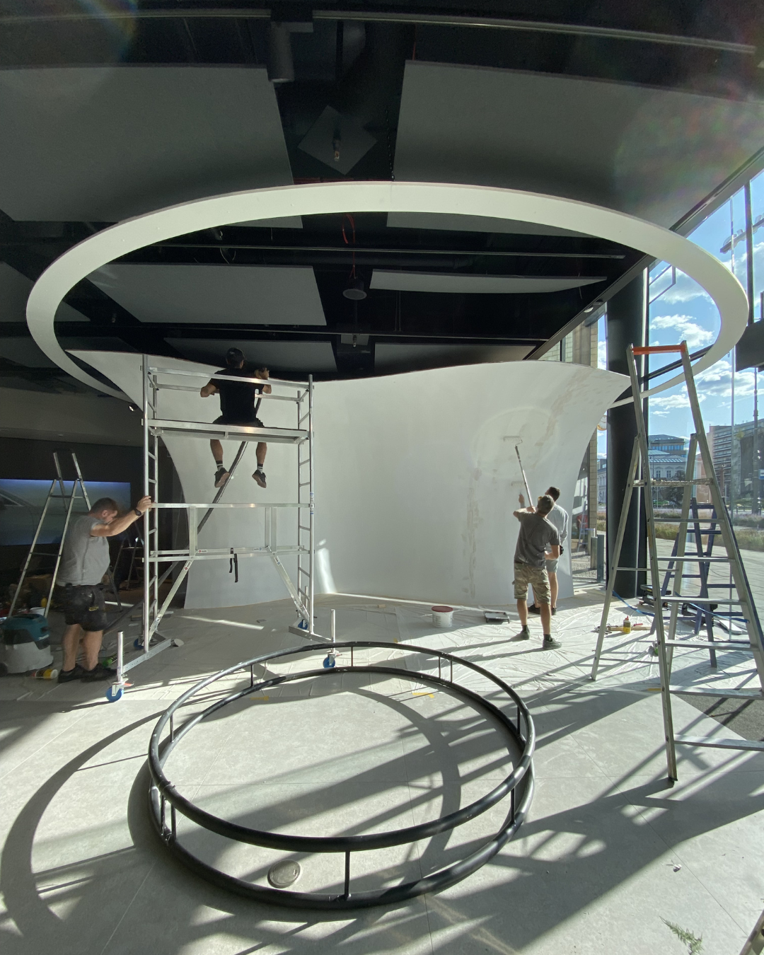 Group of workers assembling a modern indoor sculpture or installation, involving scaffolding, ladder, and tools, with a large semicircular structure and a curved wall, near a glass wall with sunlight outside.