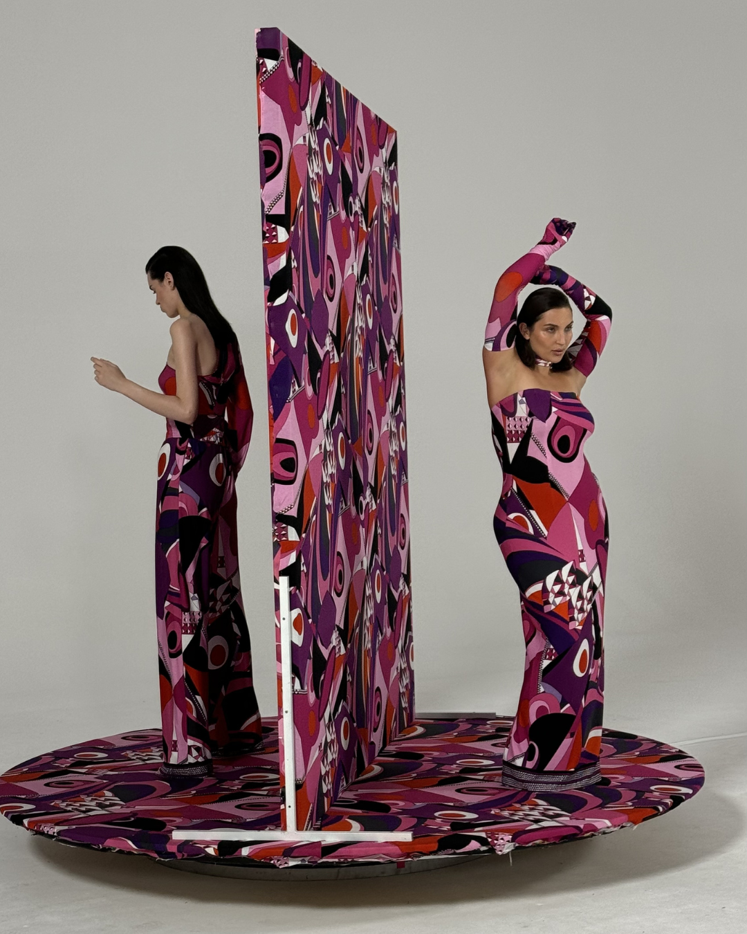 Two women in colorful, abstract patterned dresses are standing on a rotating platform with a patterned divider in the middle, against a plain background.