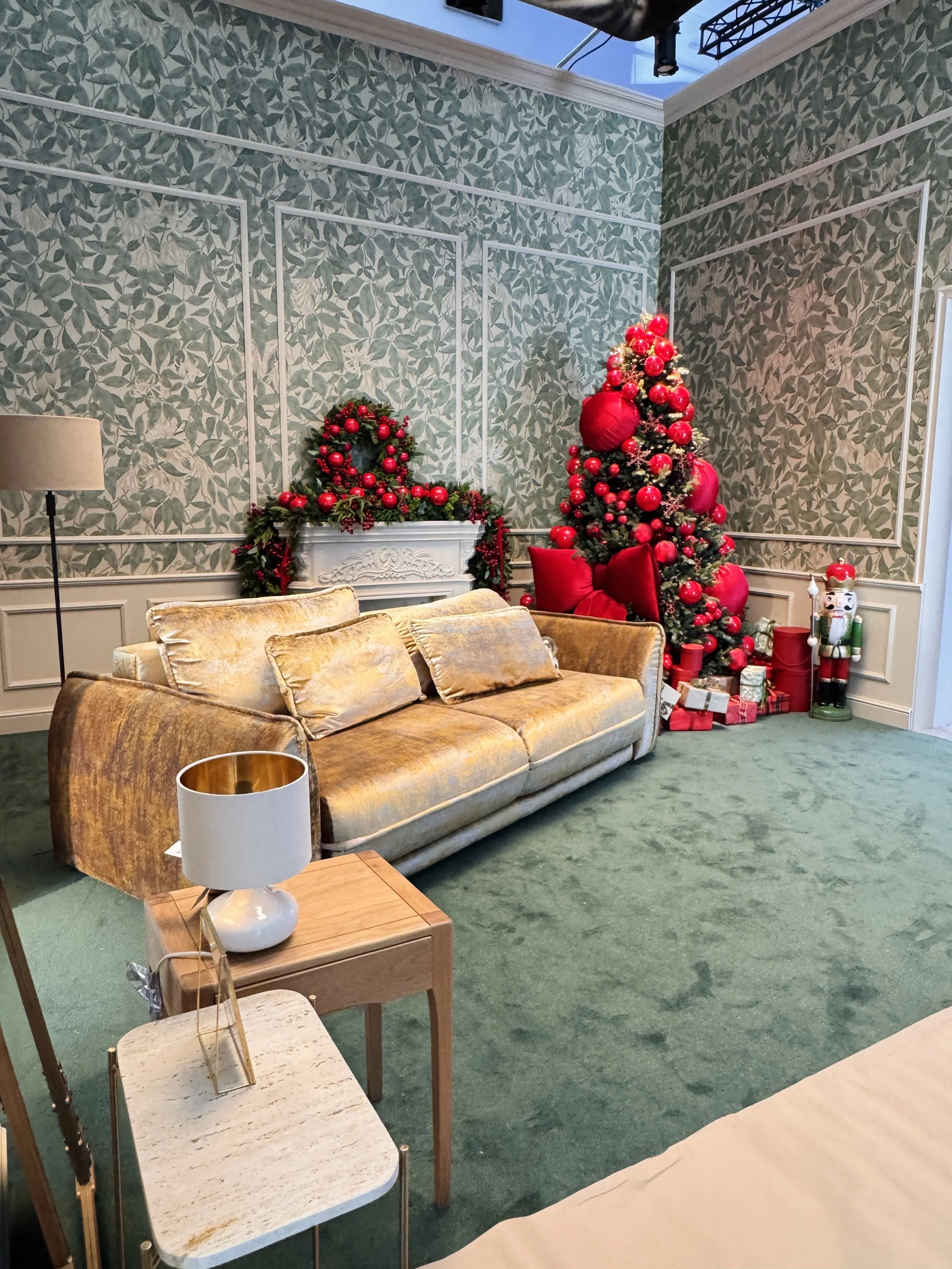 Living room decorated for Christmas with a Christmas tree, wreath on the fireplace, wrapped presents, nutcracker, and red decorations. Furniture includes a velvet sofa, side table, and lamp.