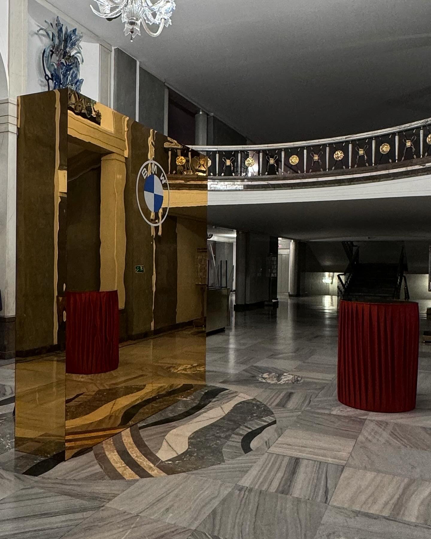 Lobby with polished marble floor, red cylindrical table, gold and black decorative panel with BMW logo, and a staircase in the background.