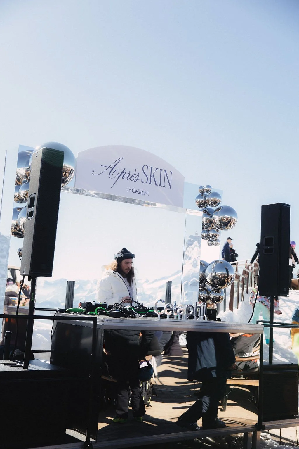 DJ performing at a snow-covered outdoor event with a backdrop of blue sky, surrounded by reflective silver balloons and black speakers, under a sign that reads 'Après SKIN by Cetaphil'.