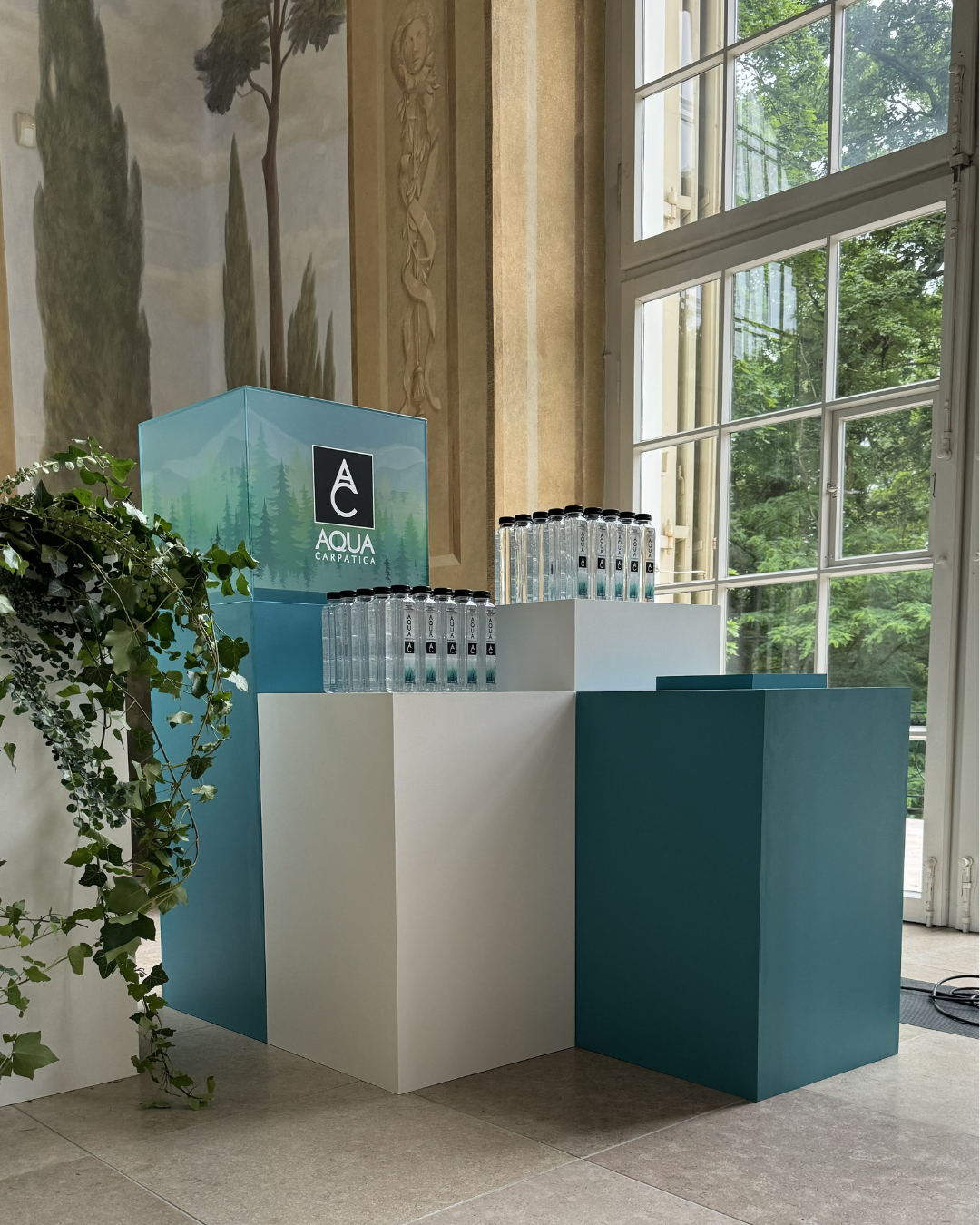 Display of Aqua Carpatica bottled water on white and blue furniture near large windows with green trees outside.