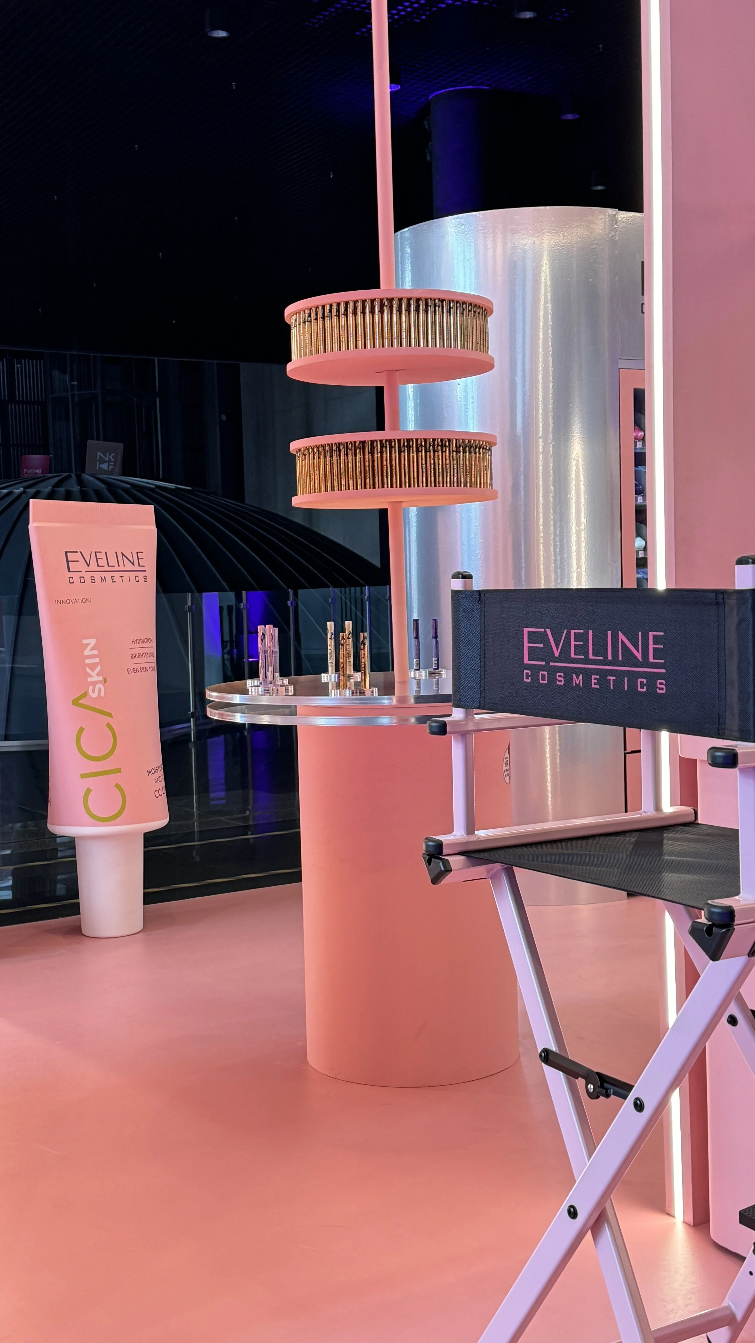 Display of Eveline Cosmetics products with a pink theme, including a large sign, lipstick tubes, and a folding chair, set against a pink and black background with subtle lighting.