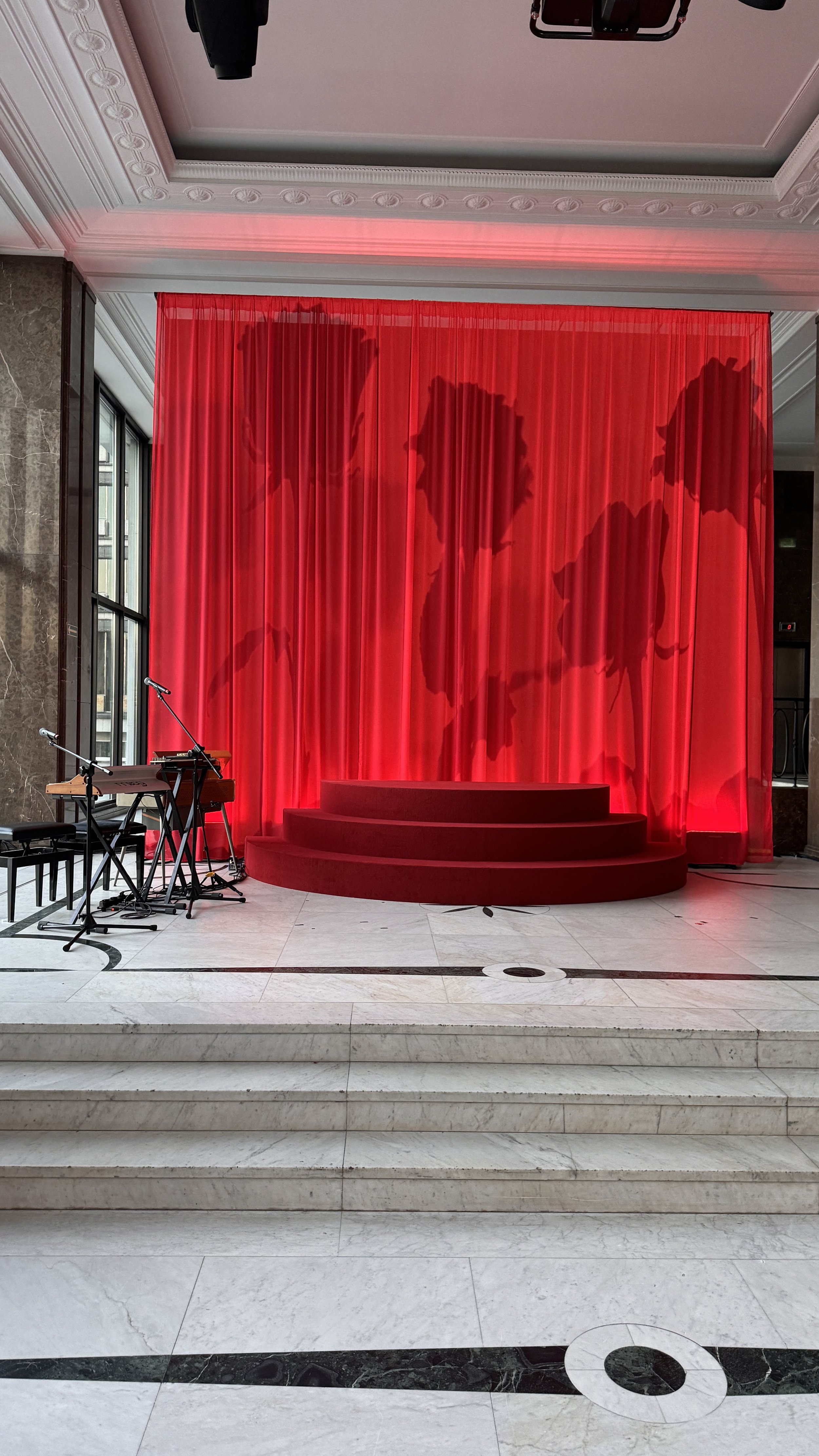 Stage with red curtains and shadows of people projected on the curtains, steps leading up to stage, musical instruments including a keyboard and microphone on the left, located in a well-lit room with large windows and marble floors.