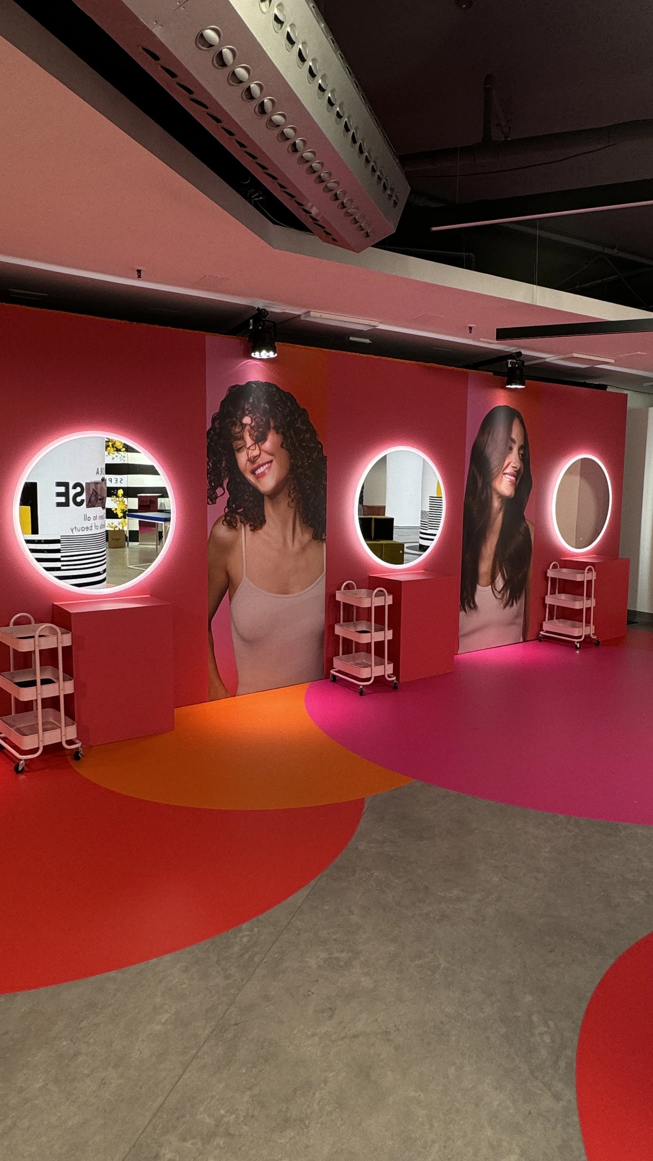 Colorful hair salon with large portraits of smiling women with curly and straight hair, pink walls with circular mirrors, and pink and orange patterned flooring.