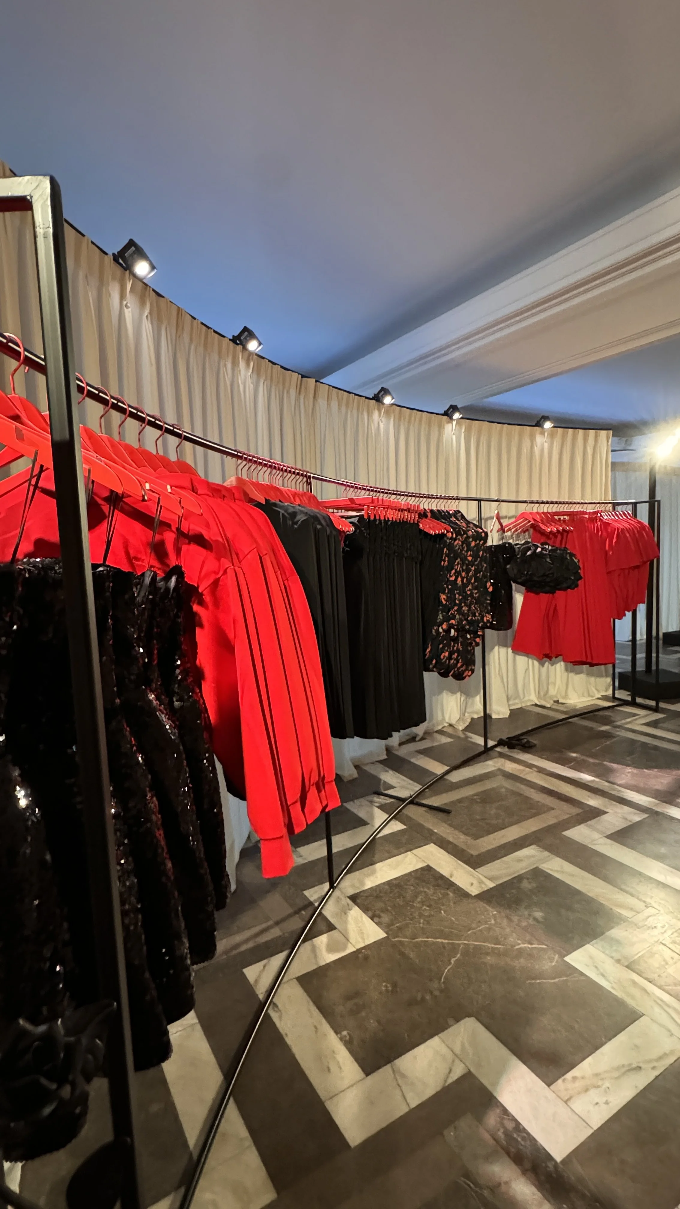 Clothing racks with red, black, and floral dresses in a boutique or fashion event, with overhead lighting and patterned marble flooring.