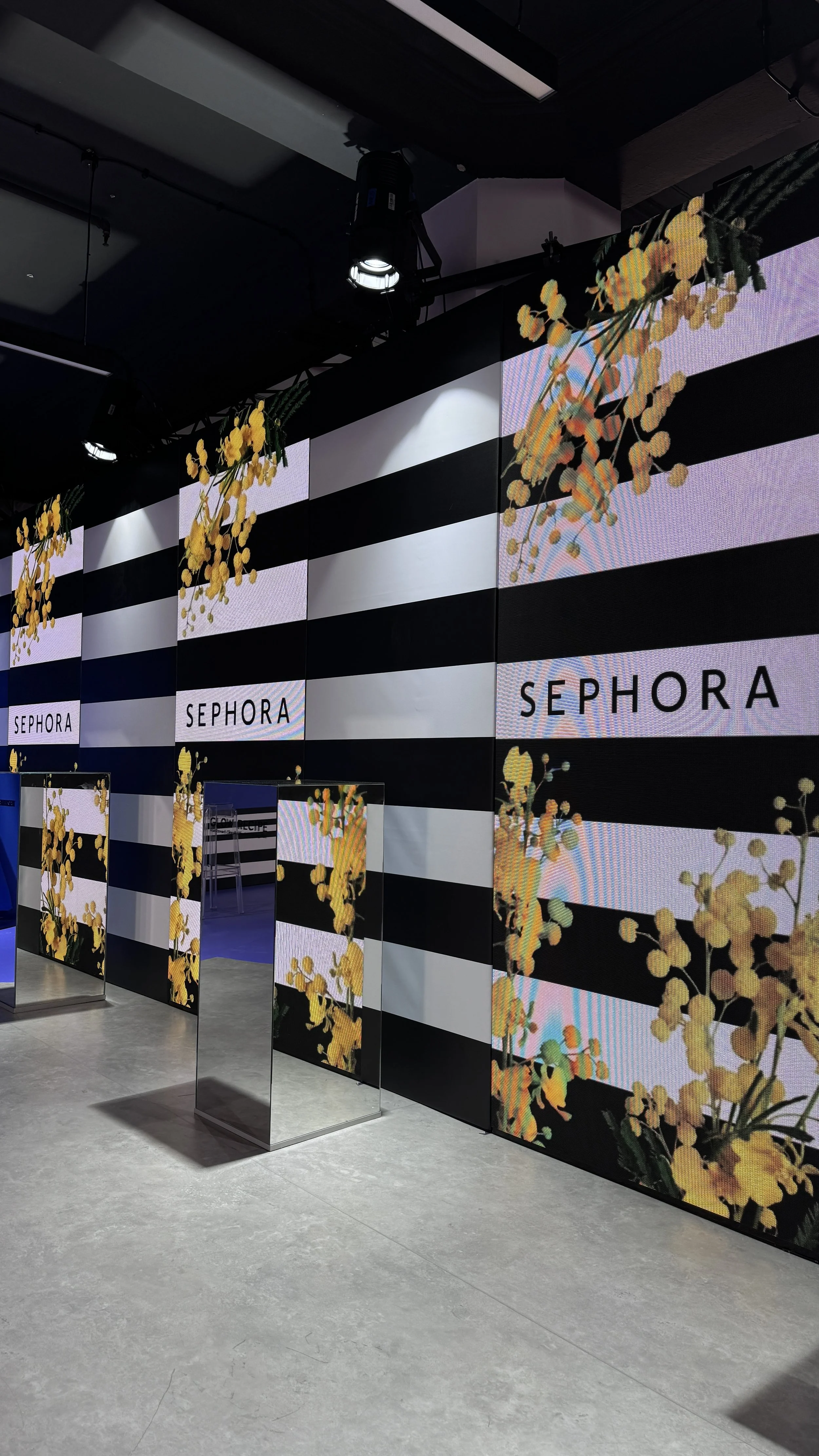 Indoor event space with black and white striped walls and digital screens displaying the Sephora logo and floral designs, with a few mirrors on stands in the foreground.