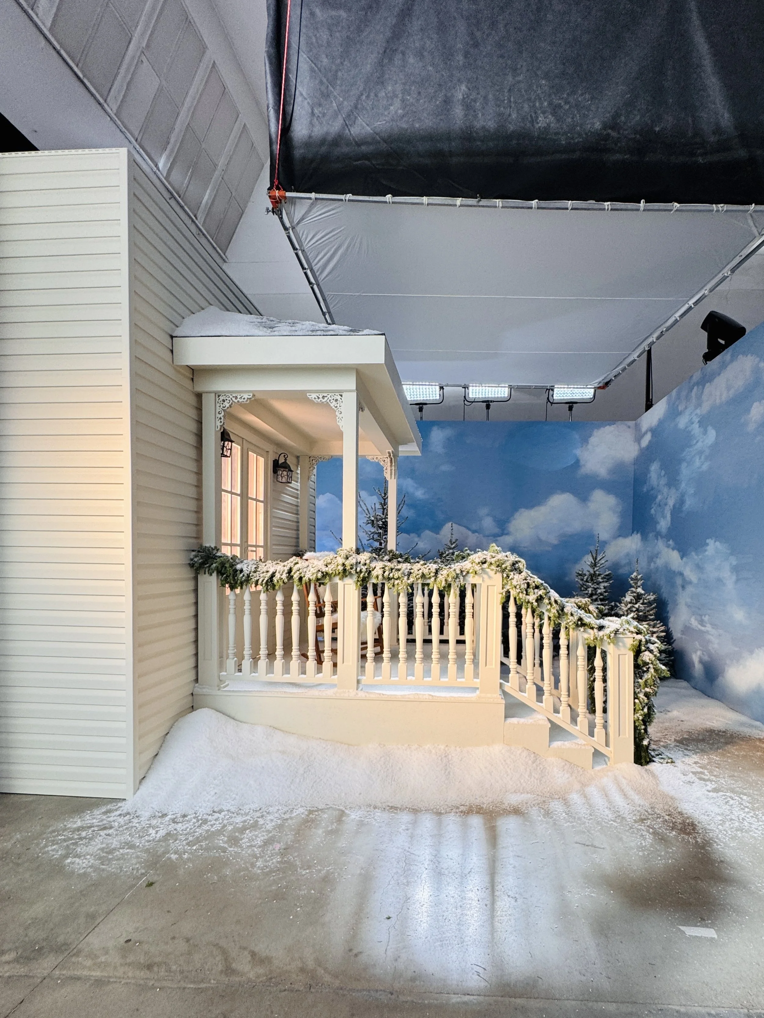 A decorative house porch with white railing, hanging flower pots, and a snow-covered roof. The background has a painted sky with clouds and snow-covered trees.