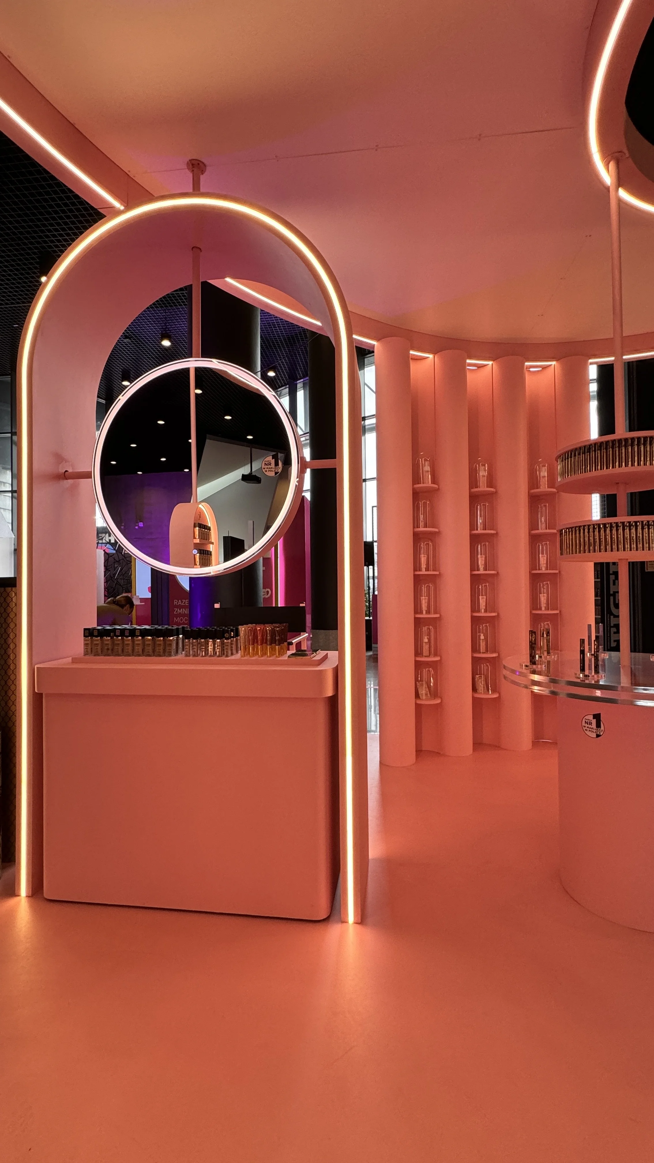 Inside a modern building with pink and orange lighting, featuring display shelves with small bottles and bottles arranged on counters, and circular architectural elements.