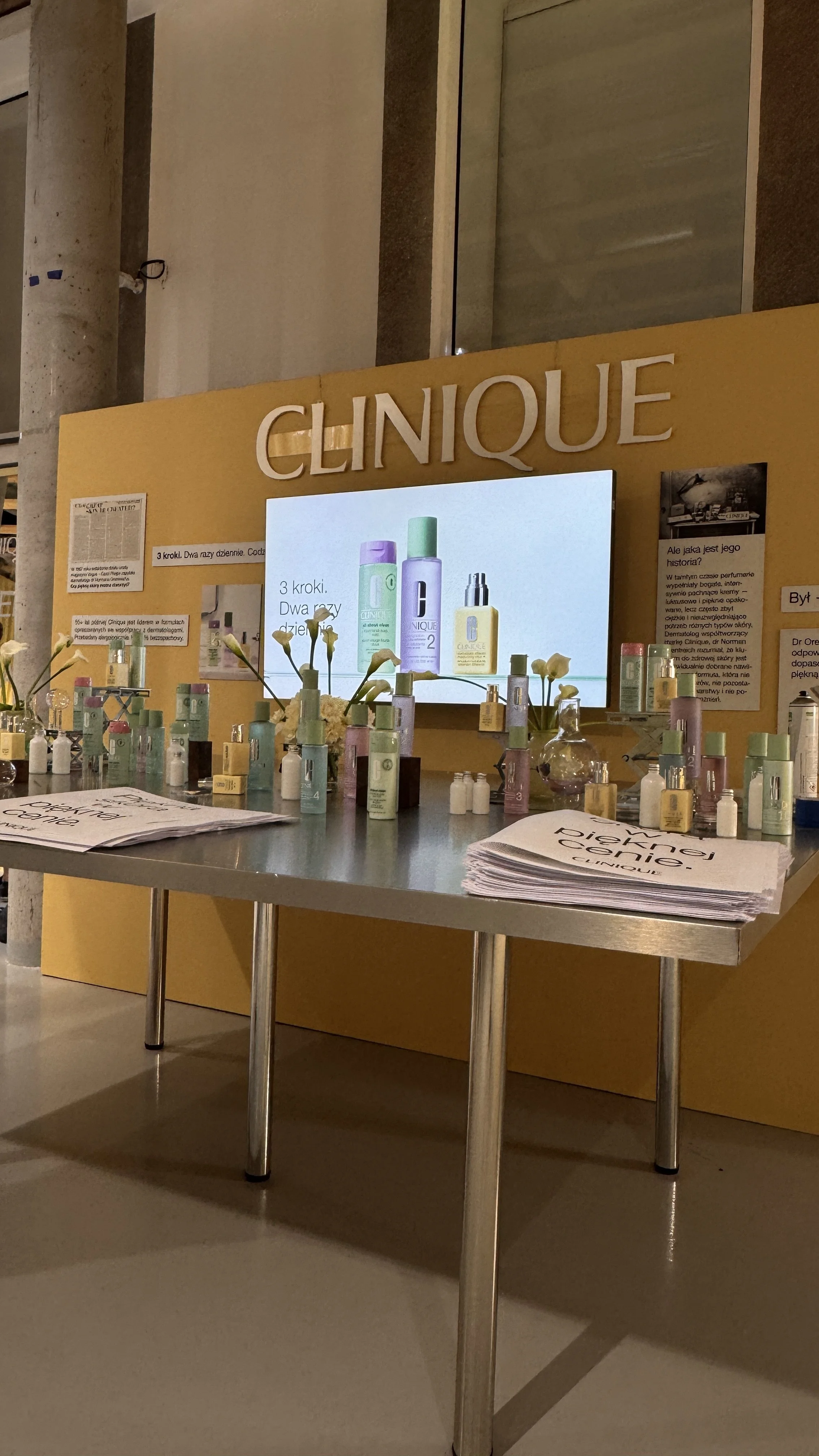 A display table with various skincare products, flowers, and informational pamphlets in front of a yellow booth with the word 'Clinique' and a digital screen showing product images and descriptions.