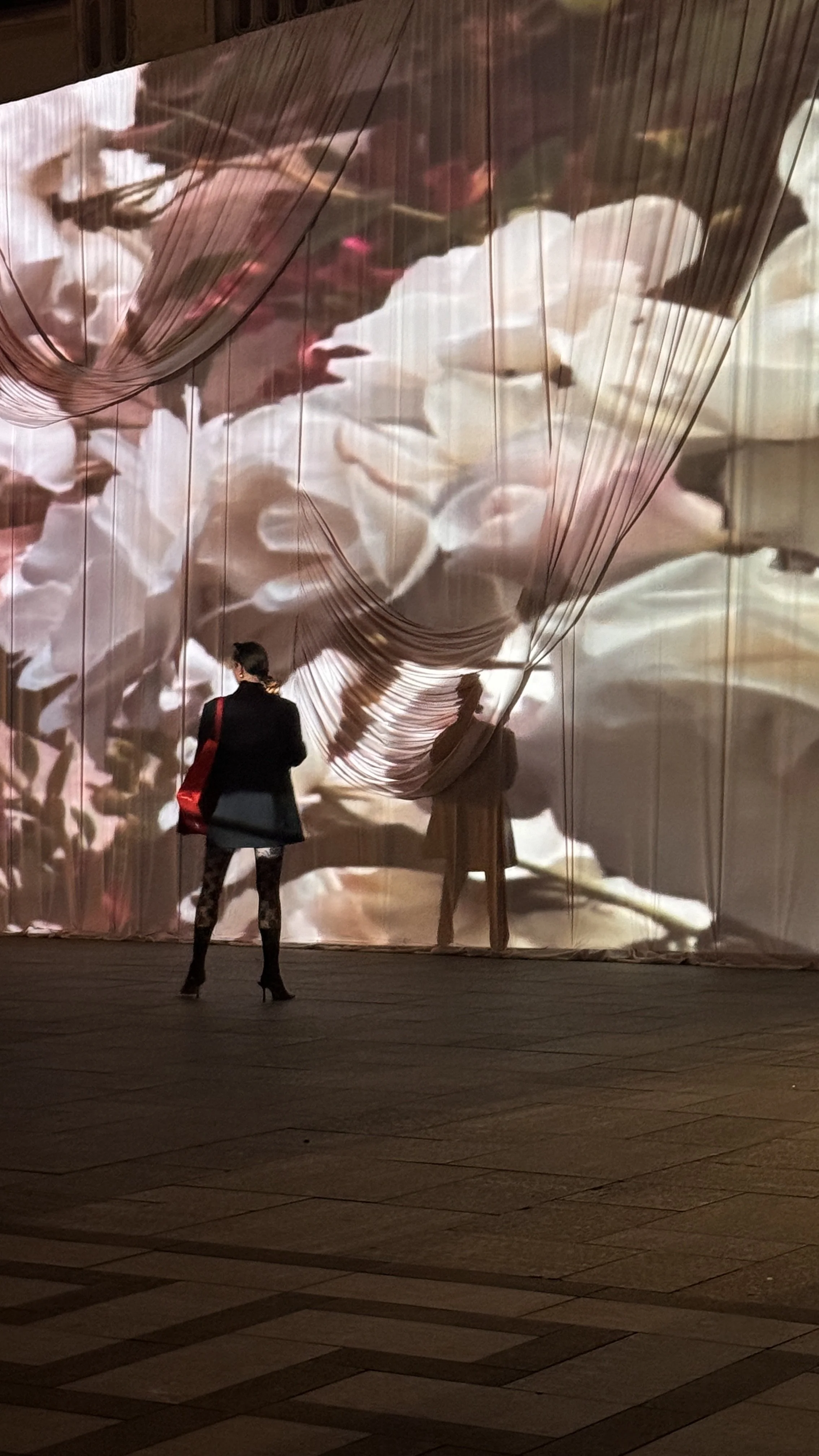 A woman with dark hair, wearing a black blazer, leggings, and high heels, stands facing a large digital display with flowing flower patterns. She has a red bag over her shoulder and is viewing the artwork on the display.