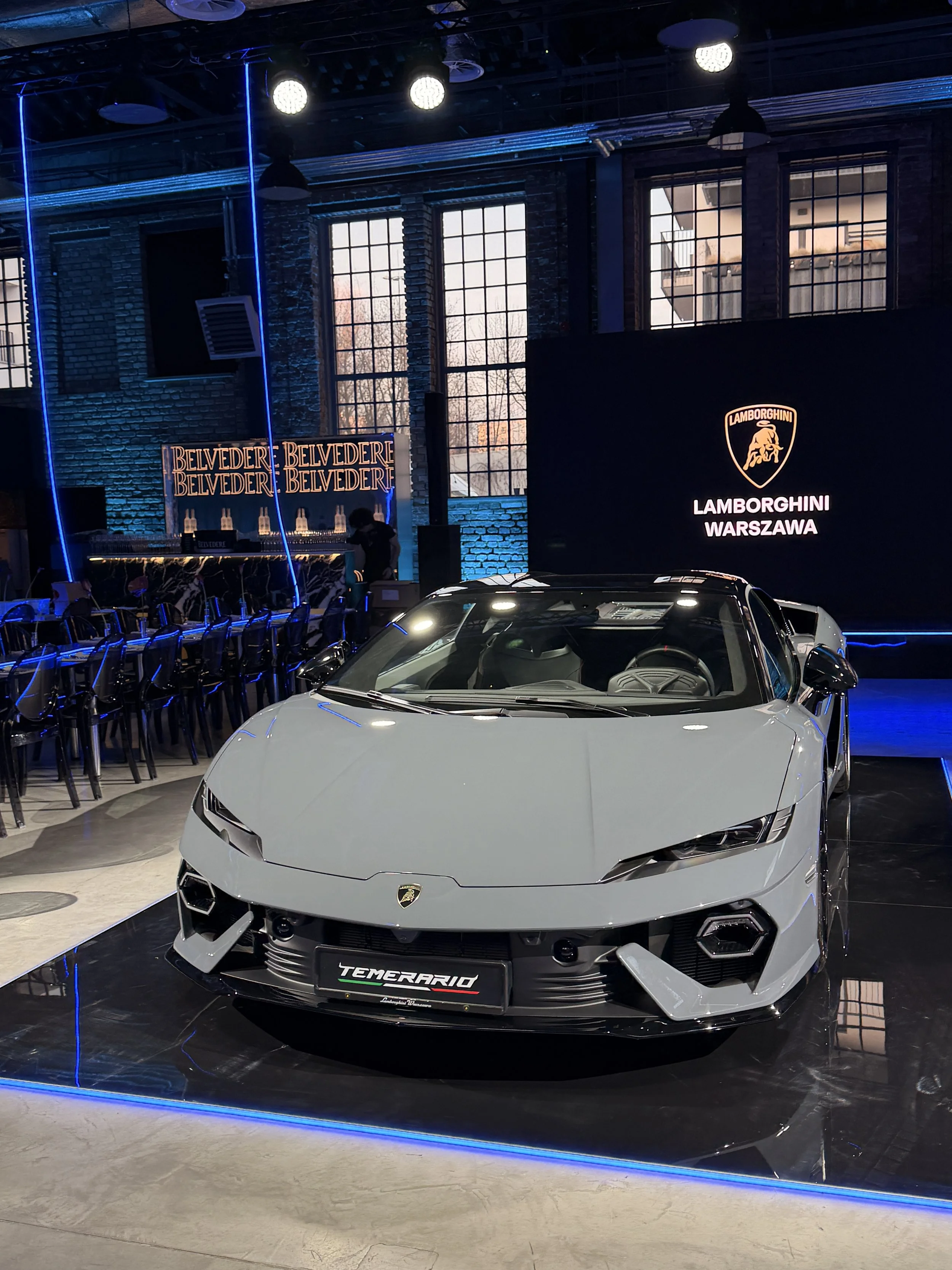 A white Lamborghini sports car on display at an indoor event with blue and yellow lighting, showing the Lamborghini logo and the model name 'Terarrio' on the front grille.