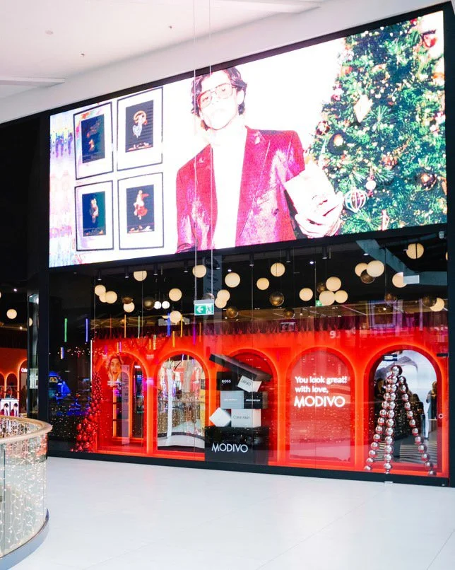 Christmas-themed window display at a mall featuring a large digital screen showing a person in a red outfit next to a decorated Christmas tree, with framed holiday artwork inside the store and festive decorations outside.