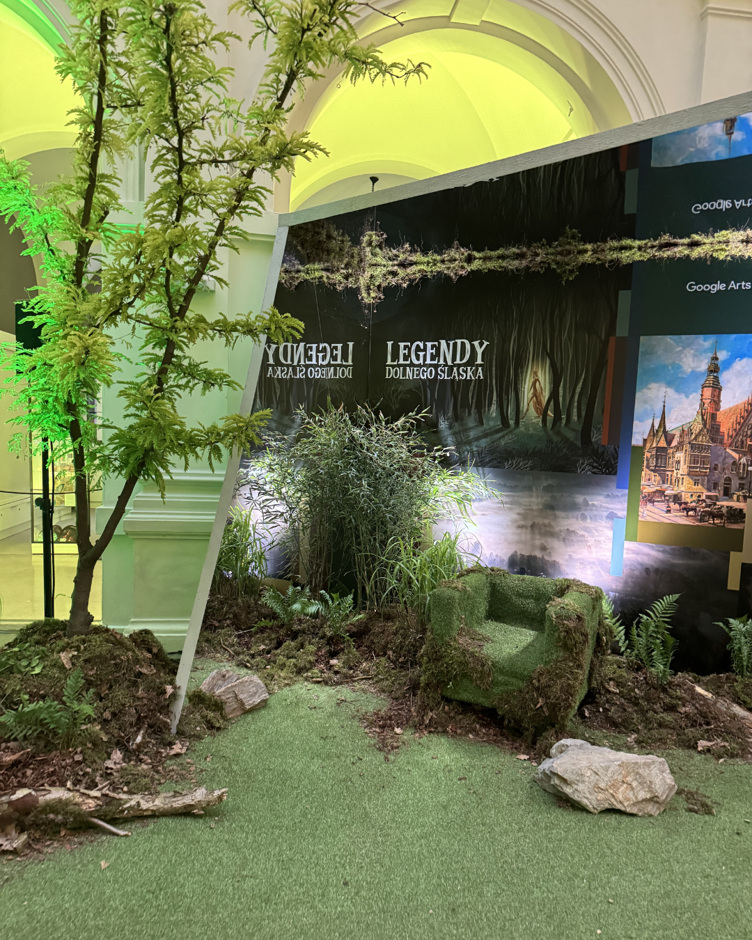 Indoor display with artificial grass, plants, a moss-covered chair, and a backdrop with forest and historic city images, promoting Legends of Lower Silesia.