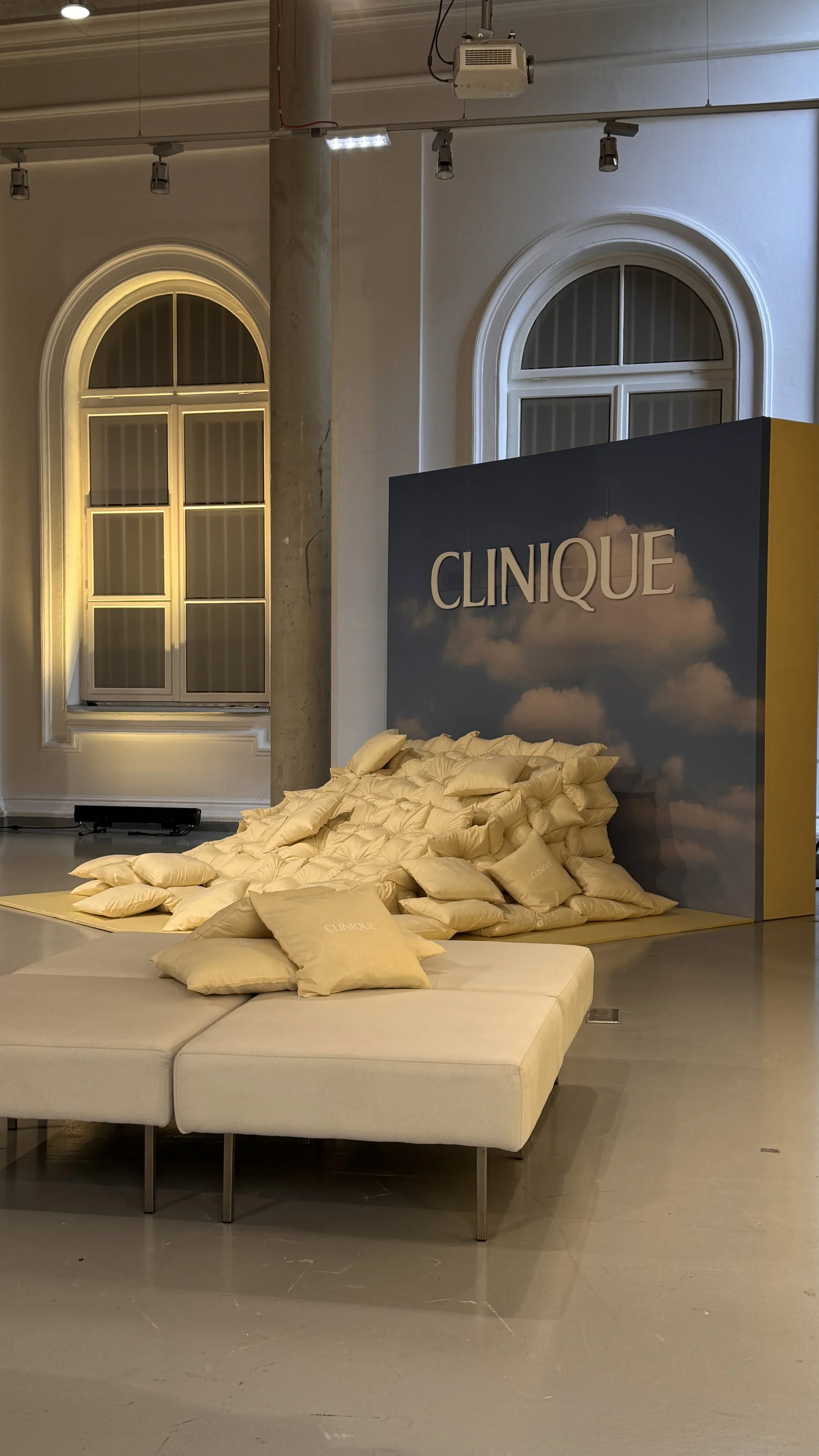 An indoor exhibition space with a display made of numerous yellow pillows and cushions arranged around a dark backdrop with 'Clinique' written on it. The room has large arched windows and is lit with overhead lighting.