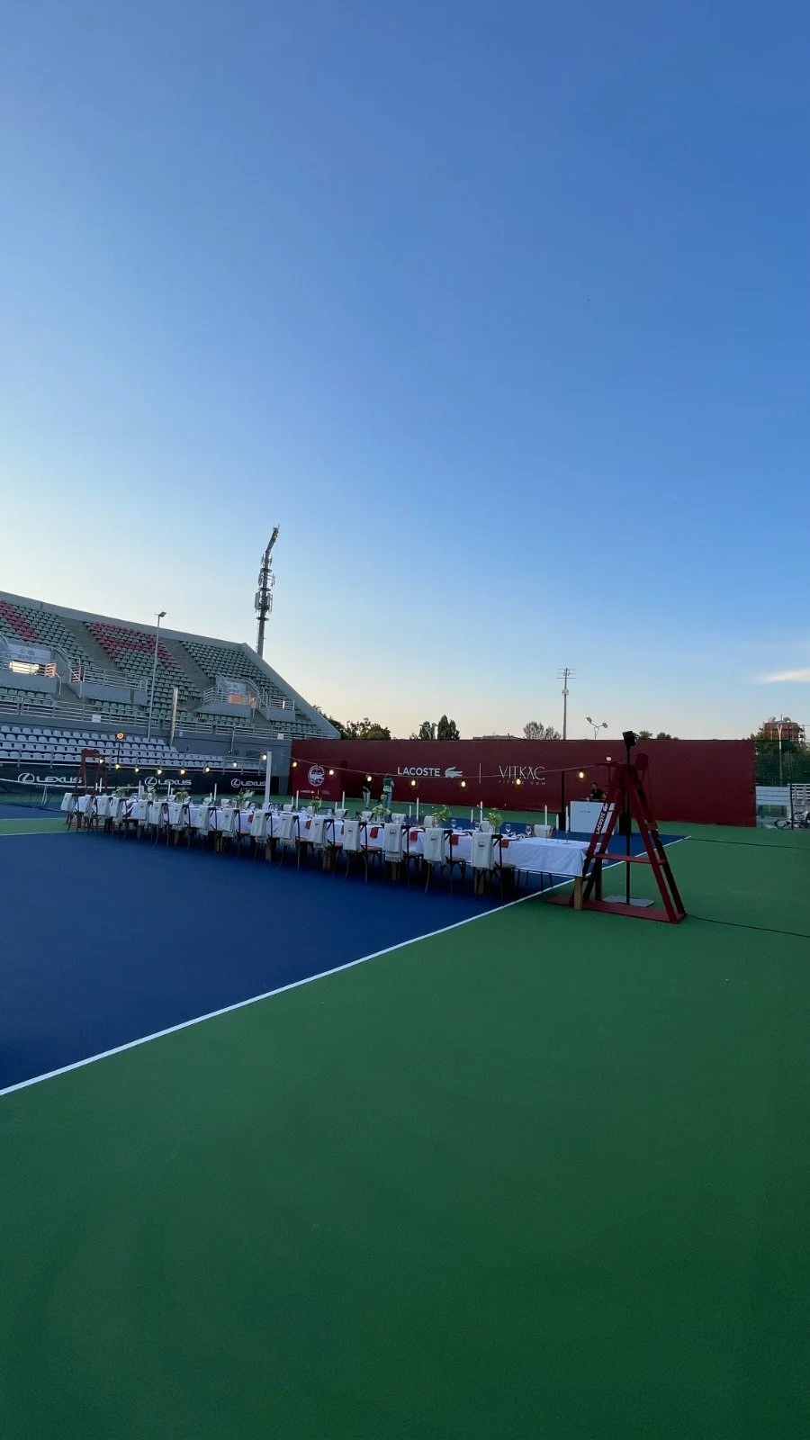 Empty tennis or sports court with a long dining table set up along the sideline, under a clear blue sky, with stadium seats and advertisement banners in the background.
