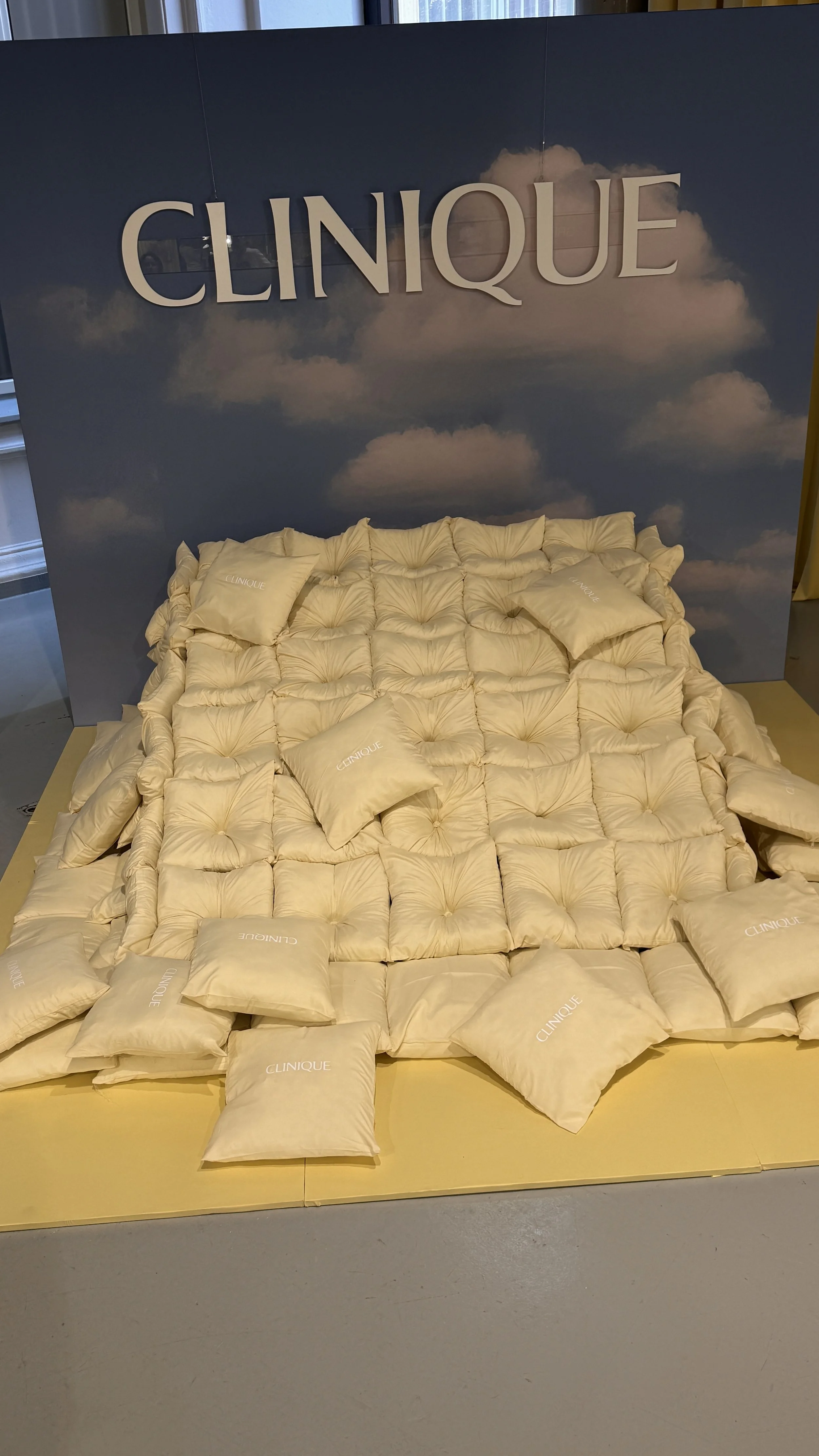 Display of plush beige pillows and bedding with the Clinique logo, set against a backdrop with the Clinique name and cloud imagery.