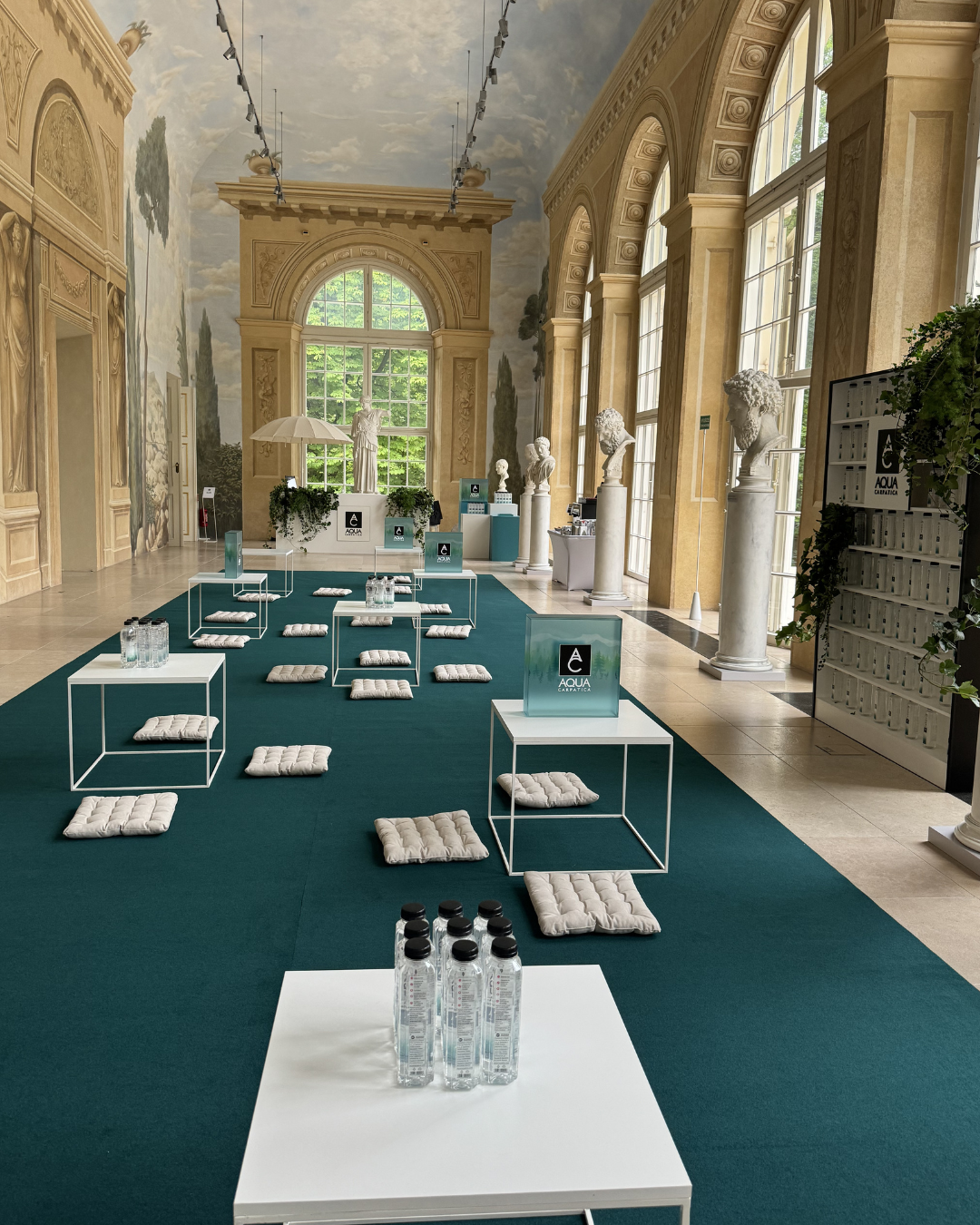 A spacious, elegant hall with large windows, classical statues, and wall murals, set up for a presentation or seminar with white tables, cushions on the floor, and water bottles.