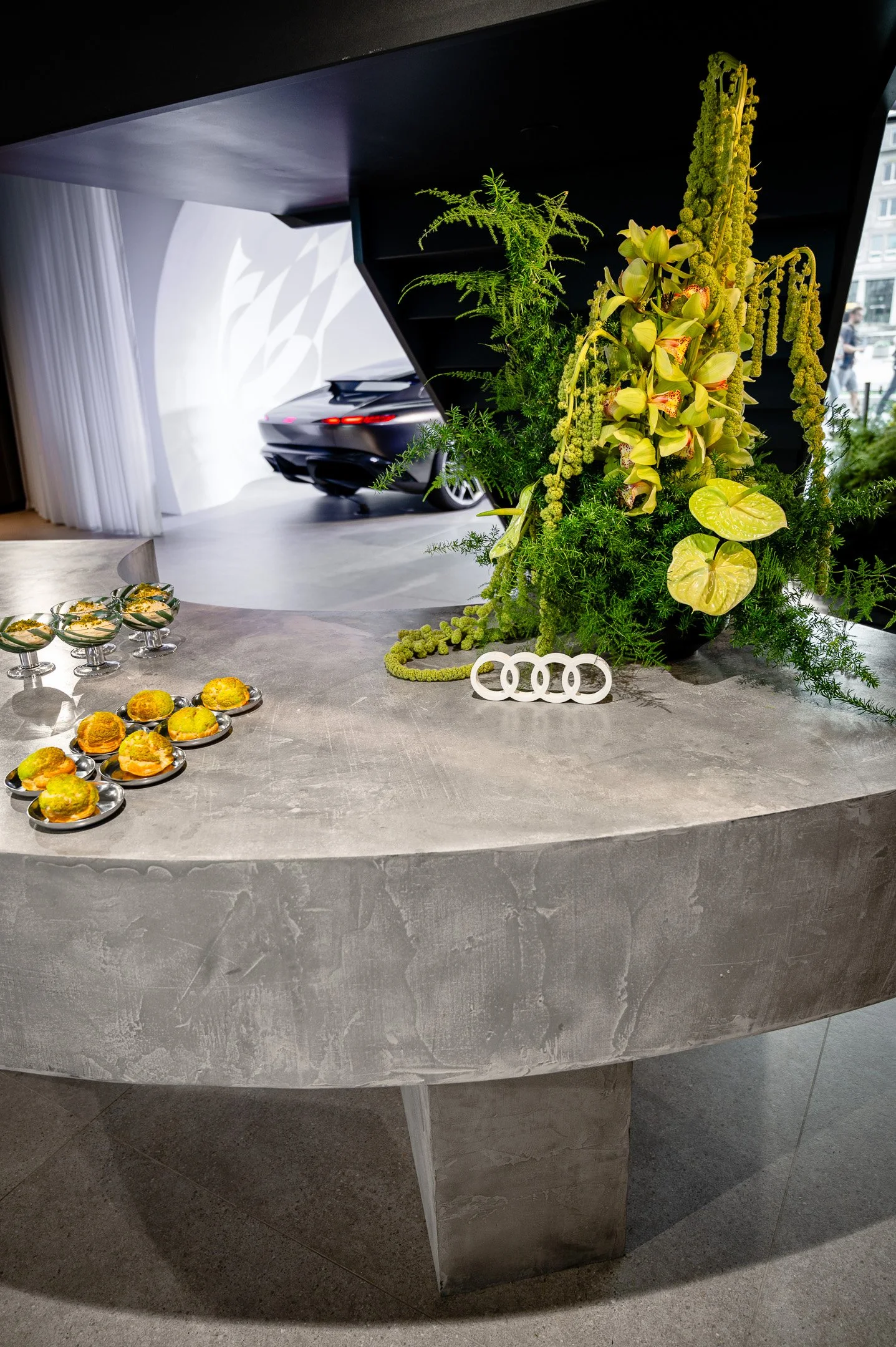 A large modern gray marble table with a floral arrangement of green and yellow flowers and foliage, and small plates with food, in an indoor space with a view of a city street outside.