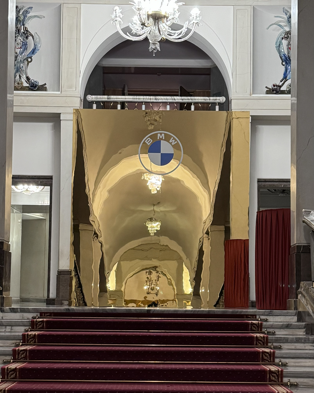 An elegant hotel lobby with a staircase covered by a red carpet, a shiny gold BMW banner with the BMW logo hanging above, ornate chandeliers, and decorative sculptures in glass cases on the sides.