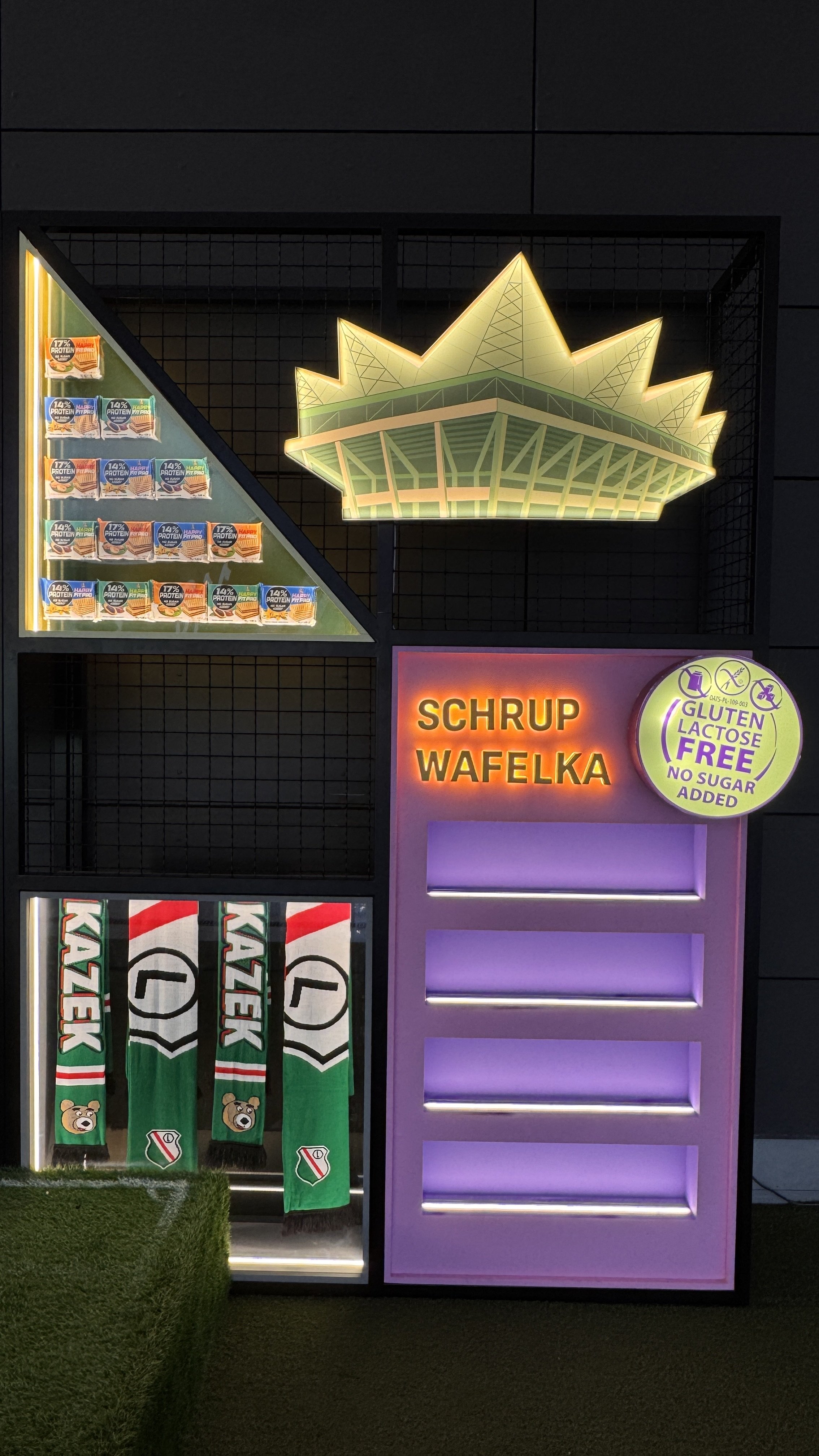 Neon sign above a food stand advertising waffles with syrup, with a menu of small snack packs on the side, and purple-lit display shelves underneath.