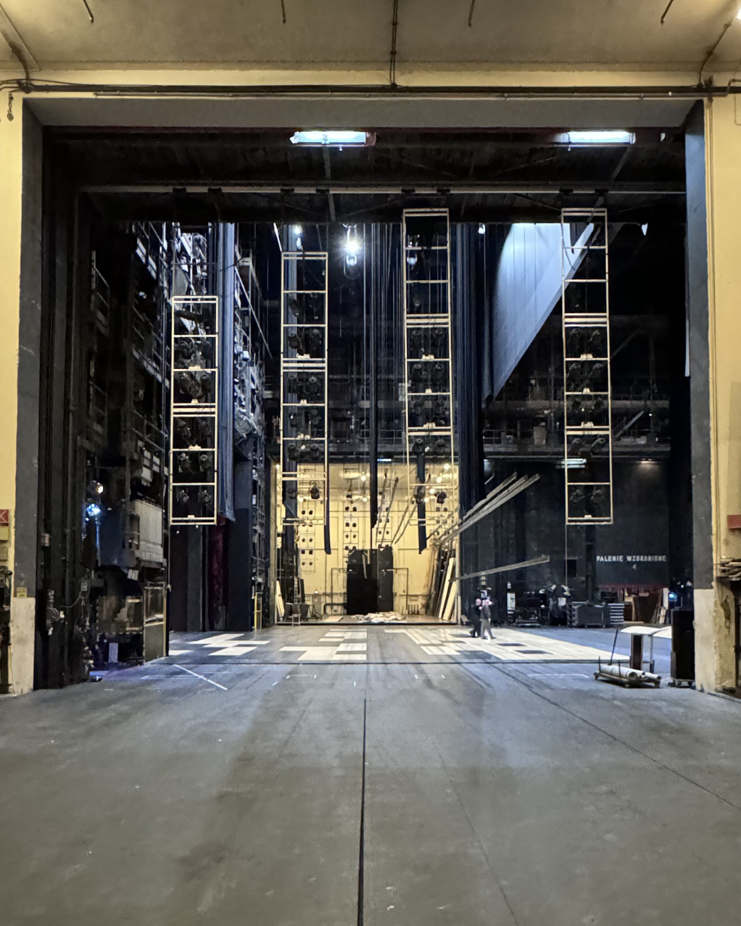 The image shows an empty theater stage with professional lighting and sound equipment hanging from the ceiling. The stage is viewed from the backstage area, with some personnel and supplies visible in the distance.