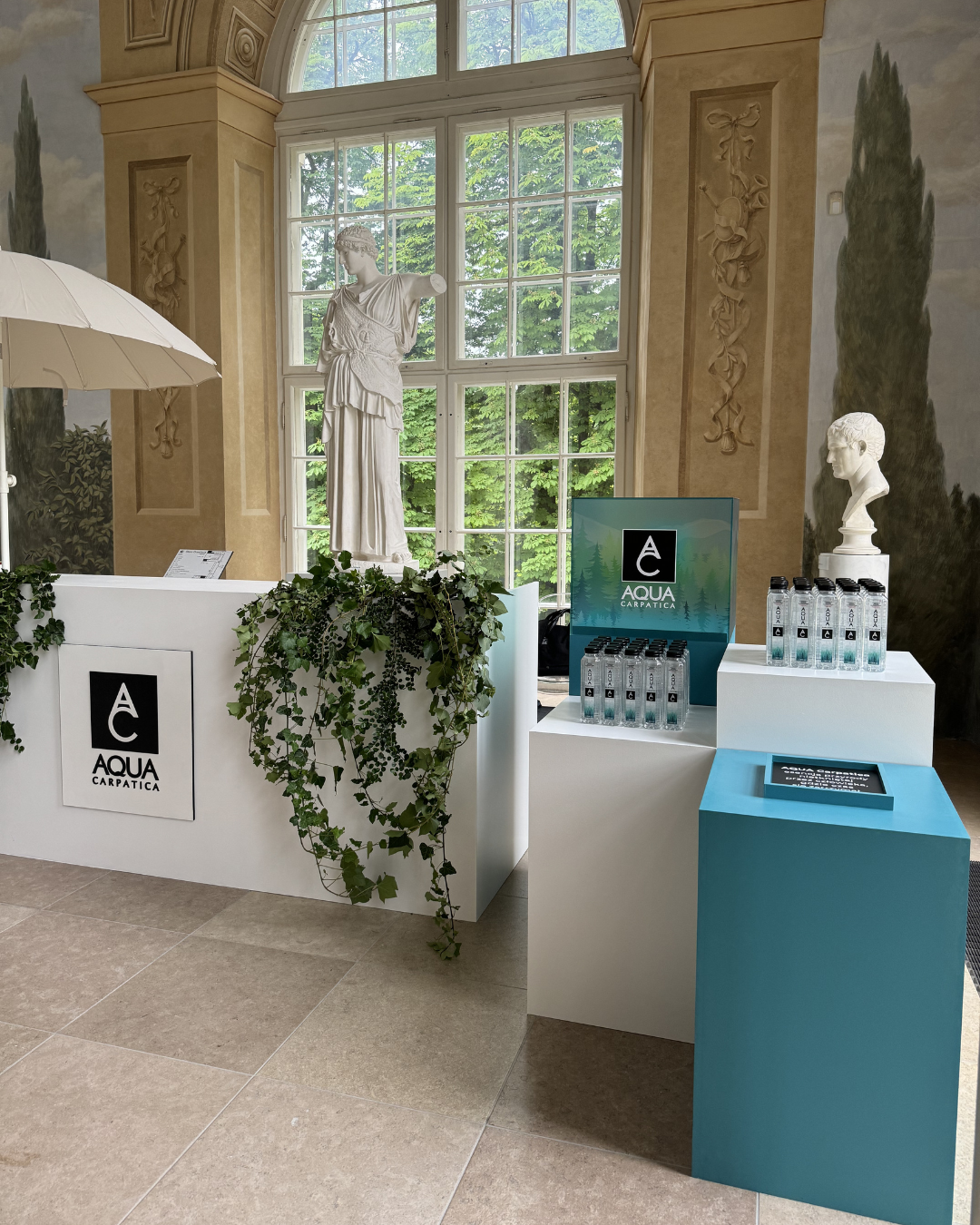 Display booth for Aqua Carpatica bottled water featuring a white counter with green ivy, a sign with the logo, and multiple water bottles arranged on white and blue stands. Two classical sculptures and a large window with greenery outside add an eleg
