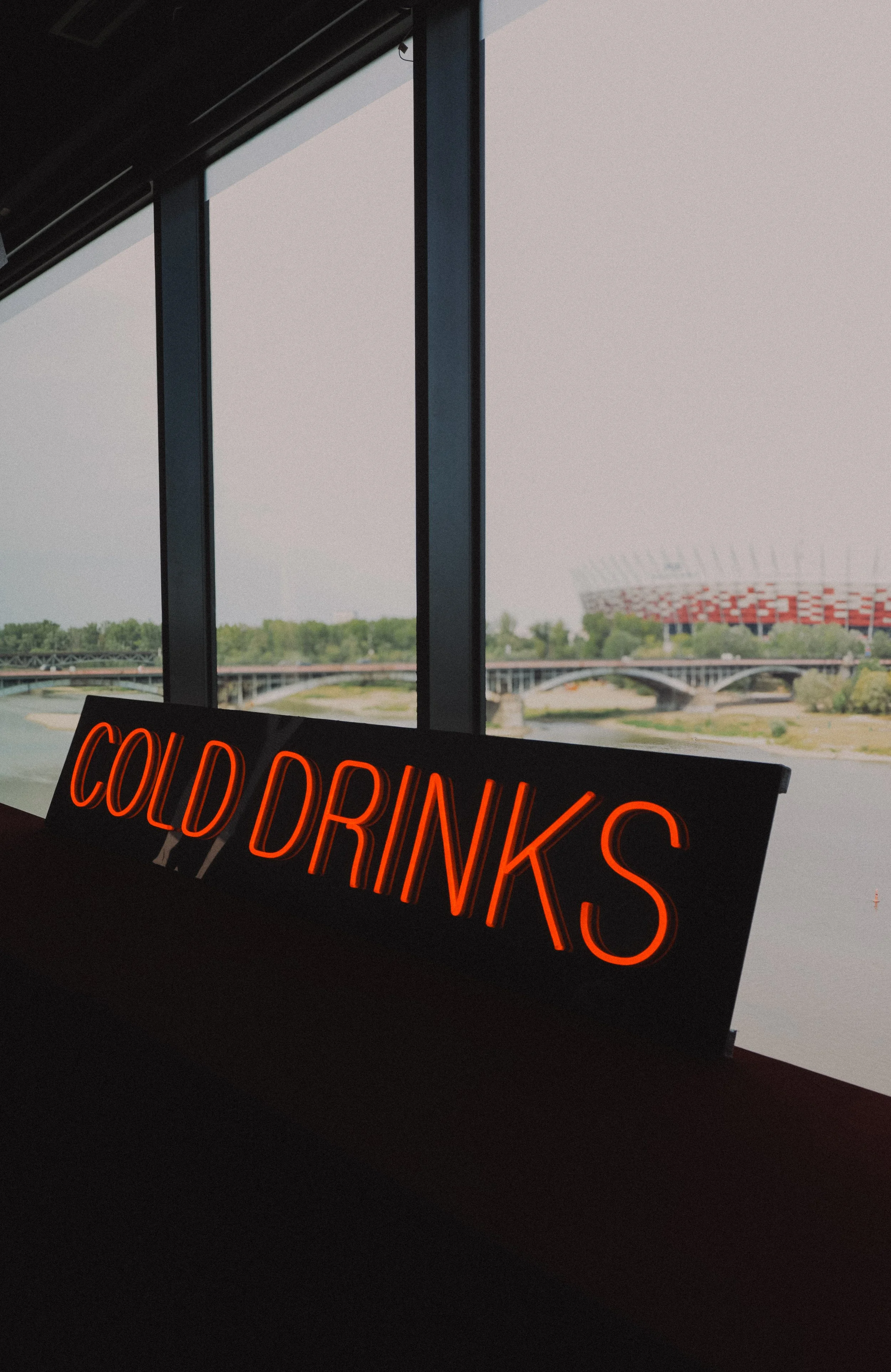 View from inside a building showing large windows, a neon sign that says 'Cold Drinks', and a distant view of a stadium and a bridge outside.
